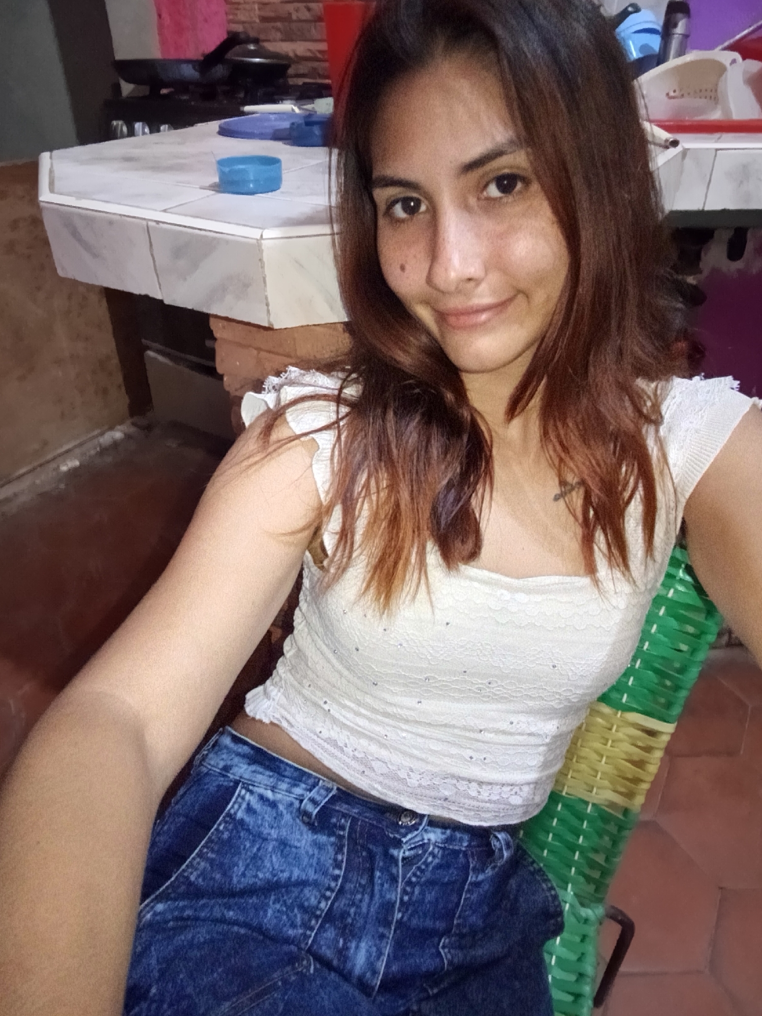 In Juan Griego, Venezuela, a young woman with long, brown hair and light skin sits on a chair made of woven green and yellow plastic. She is wearing a white lace crop top with sparkling embellishments and high-waisted, acid-washed denim shorts. The woman is looking at the camera and smiling. In the background, a tiled countertop with blue containers and a stovetop with cookware are visible. The floor is made of reddish-brown tiles.