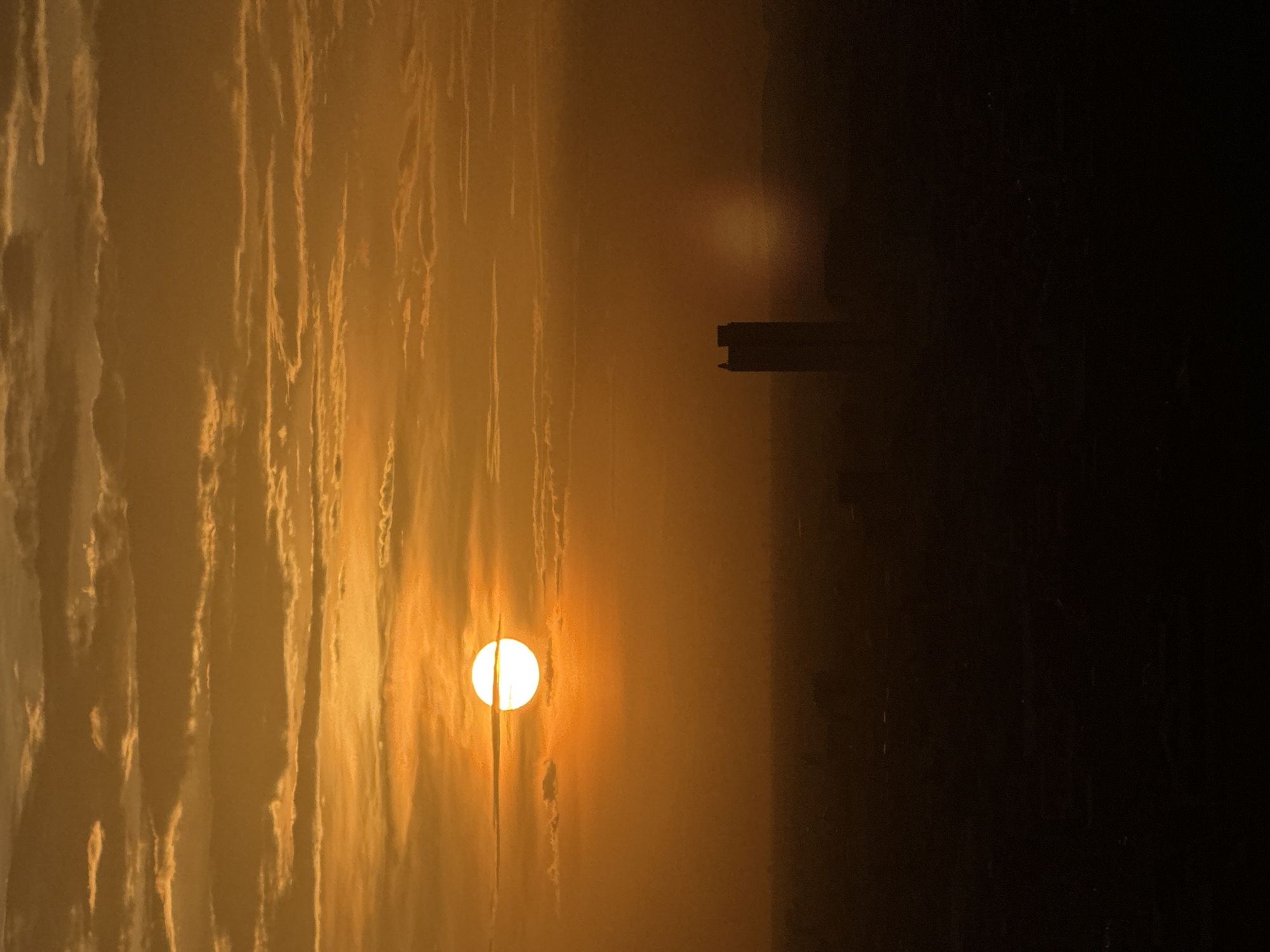 The image captures a sunset over a city, with the sun a bright, glowing orb partially obscured by horizontal clouds. The sky is a warm gradient of orange and yellow. In the middle ground, a silhouetted skyscraper stands prominently against the hazy atmosphere. The foreground depicts a dark, silhouetted cityscape, suggesting a vast urban expanse. The scene is one of urban twilight, with a single tall building dominating the distant view. Based on the provided information, the location is the city of Khan Mean Chey, Cambodia.