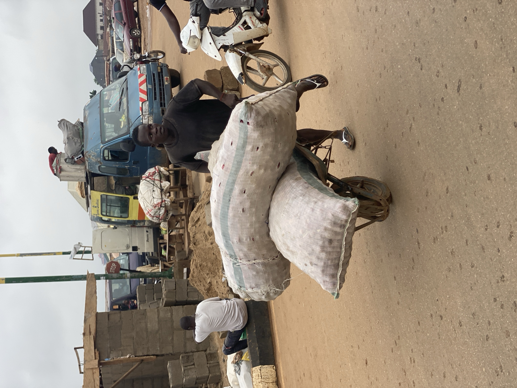 The image shows a man in Kafanchan B, Nigeria, carrying two large sacks on a bicycle. The man is wearing a black shirt and sandals. The sacks appear to be filled with white produce, possibly onions or potatoes. The bicycle is old and rusty.
In the background, there are several vehicles, including a blue truck with people on top and a few other smaller cars and vans. A motorcycle is parked nearby. On the left, there's a partially constructed building made of gray concrete blocks and a "Stop" sign. Another man wearing a white shirt is kneeling near the building. The ground is paved, showing signs of wear and tear. The sky is overcast, suggesting a cloudy day.