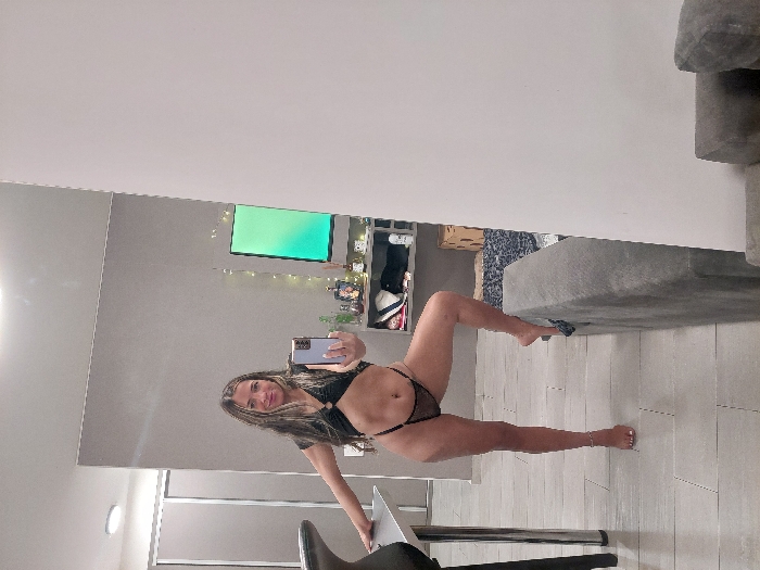 An adult female is depicted in a residential interior in Buenos Aires, Argentina, taking a mirror selfie. She has long, dark hair and is wearing a black two-piece swimsuit or lingerie set, consisting of a crop top and high-cut thong bottoms. Her left foot, with white-painted toenails and an ankle bracelet, is bare on a light grey tiled floor. Her right bare foot is elevated and resting on the armrest of a grey fabric sofa, which occupies the lower right foreground. She holds a light-colored smartphone in her right hand, angled towards the unseen mirror, and smiles directly into the camera's reflection. Her left hand rests on the chrome leg of a light-colored table to her left. The background features a plain grey wall on the left transitioning to a white wall on the right. A wall-mounted television displaying a bright green image is visible in the mid-background, above a shelf adorned with various bottles, small plants, and string lights. A patterned blue and white rug is partially visible under the sofa and beneath the shelf. A closet with light-colored sliding doors is seen in the far left background, and a cardboard box rests on the rug behind the sofa.