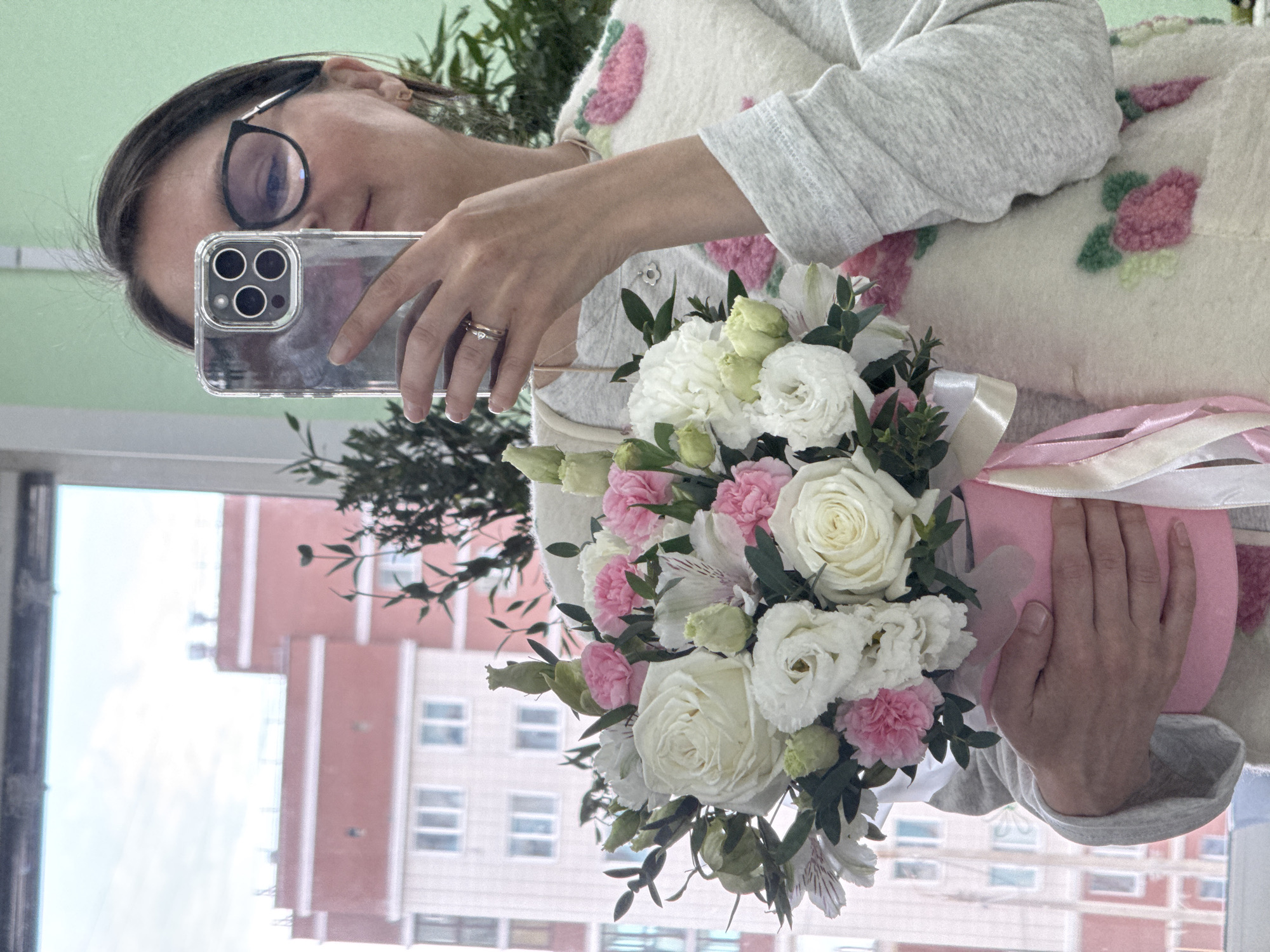 A woman is depicted taking a mirror selfie, holding a bouquet of flowers, likely indoors in Izhevsk, Russia. She is seen from the chest up, wearing dark-rimmed glasses and a light-colored long-sleeved sweater with a white garment underneath that features pink and green floral embroidery. Her dark hair is visible around her face.
She holds a smartphone with a clear case and a triple-lens camera system, which partially obscures her face. A ring, possibly a wedding or engagement ring, is visible on her left ring finger. In her other hand, she holds a lush bouquet of flowers, predominantly featuring white roses, white and pink eustoma (lisianthus), and pink carnations (dianthus), accented with various green foliage including alstroemeria. The bouquet is wrapped in pink material and tied with white and pink ribbons.
The background reveals a window reflecting outdoor urban scenery, with buildings featuring reddish-brown and white facades and numerous windows, typical of city architecture in daylight conditions. A light green wall or surface is visible in the upper part of the reflection. Some green plant foliage is also present, suggesting an indoor plant. The scene is well-lit by natural light. The image captures a personal moment, possibly celebrating the receipt of the flowers.