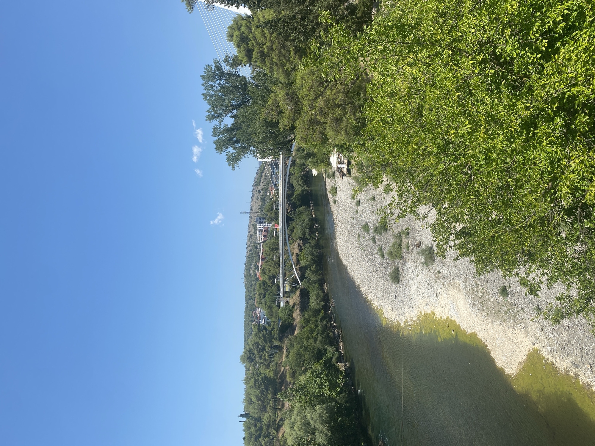 The image captures a serene view of the Morača River flowing through Podgorica, Montenegro. In the mid-ground, a modern, white cable-stayed bridge spans the river, connecting two parts of the city. The riverbanks are lined with lush green trees and vegetation, with a visible pebble beach on the right side of the river. In the background, residential buildings and structures are nestled amidst the rolling hills, indicating the urban landscape of Podgorica. The sky is a clear, bright blue, suggesting a sunny day. The scene appears peaceful and natural, highlighting the integration of urban life with the surrounding natural environment.