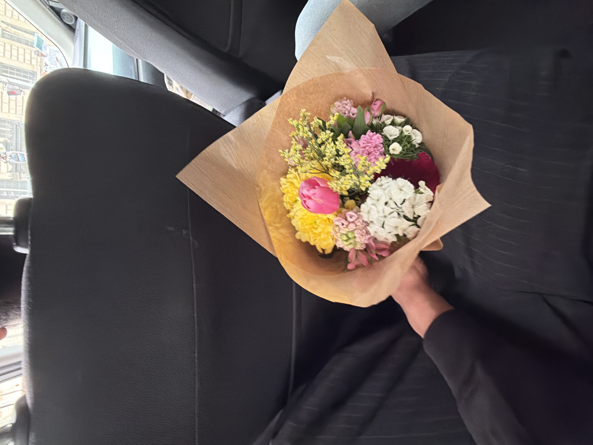 The image shows a bouquet of flowers held in a car. The bouquet is wrapped in brown paper and contains pink, yellow, and white flowers. The car has black leather seats and the person holding the flowers is wearing a black sleeve. The view from the car window shows buildings, potentially locating the scene in an urban setting. While the specific city cannot be forensically determined, the request mentions Paris, France; therefore, the scene is likely within that city.