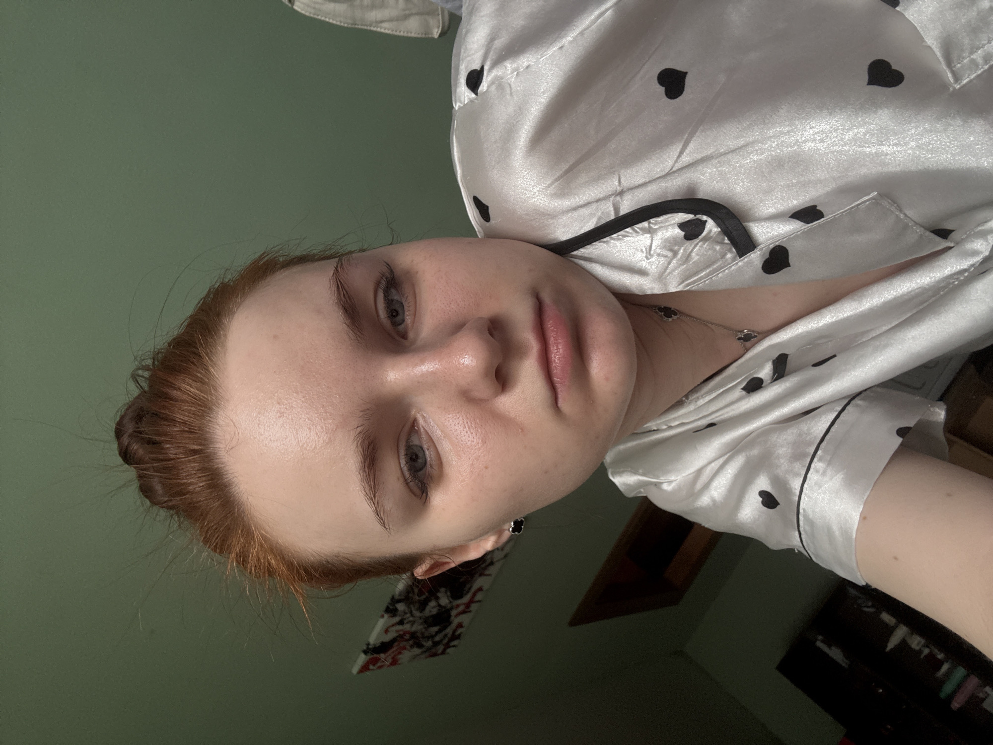 A selfie of a fair-skinned person with red hair pulled back in a bun. They have blue eyes, and their face is slightly oily. The person is wearing white satin pajamas with black hearts. The background is a blurred green wall and what appears to be a room interior. The location in Sinyavino, Russia, cannot be determined from the visual information provided.