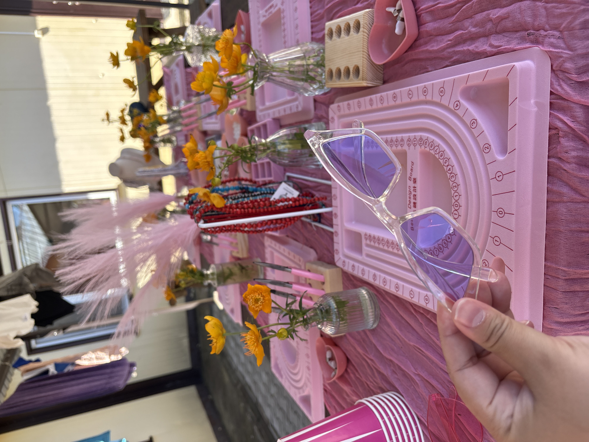 A person's hand holds a pair of clear, heart-shaped sunglasses with purple lenses over a pink bead design board. The board is on a pink tablecloth, as are several other crafting supplies, including pink cups stacked on the left, small pink heart-shaped dishes with beads, and a wooden block with holes. In the background, a table is set up with more pink bead design boards, vases of yellow flowers, and strands of red and blue beads. The scene appears to be outdoors, with a building or structure visible in the background. The image is cropped from the perspective of someone looking down at the table.
