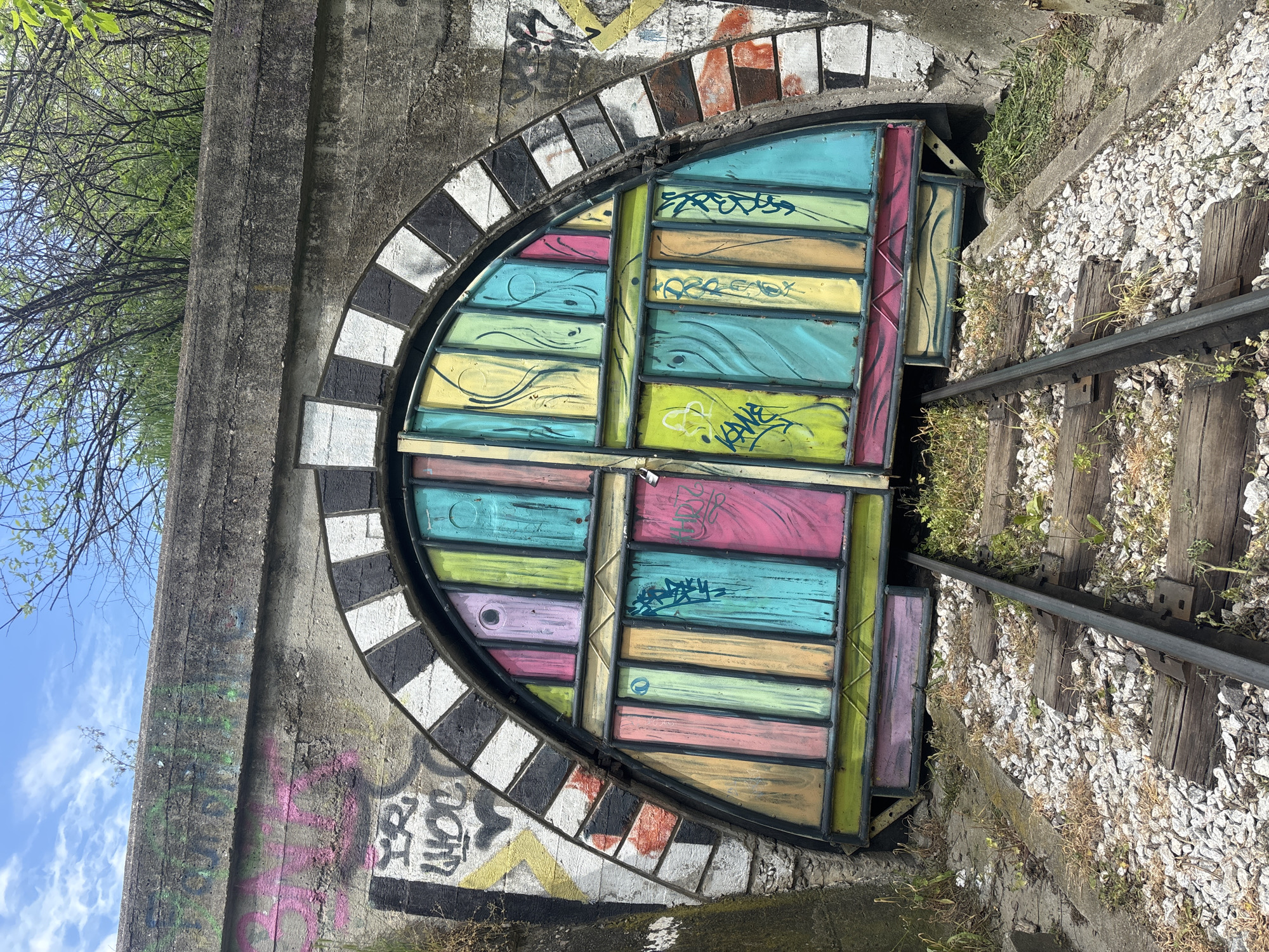 The image captures a vibrant, multi-colored barrier blocking the entrance to a concrete railway tunnel, set outdoors under bright daylight with a blue sky and sparse white clouds visible. The arched tunnel entrance is made of weathered, grey concrete. A striking black and white checkered pattern outlines the inner rim of the archway, appearing to be painted onto individual bricks or tiles.
The barrier itself is a double-door gate, constructed from horizontal wooden planks or panels, each painted in a distinct pastel hue such as pink, teal, lime green, yellow, and light purple. These colorful panels are extensively covered with black and white graffiti tags and stylized squiggles. Visible text on the gate includes "FREEZ" on a blue panel, "KANE" on a yellow panel, and "YAM" on another yellow section.
Two old railway tracks lead into the tunnel, featuring wooden sleepers laid on white gravel ballast. The tracks are overgrown with weeds and dry grass, indicating disuse. The surrounding concrete walls of the tunnel entrance are also heavily adorned with various graffiti tags in black, pink, yellow, and orange, with partially visible text such as "ZOM" and "DON'T." In the upper left background, tree branches with light green foliage are visible against the sky. The scene depicts a blend of urban infrastructure and street art in Plovdiv, Bulgaria.