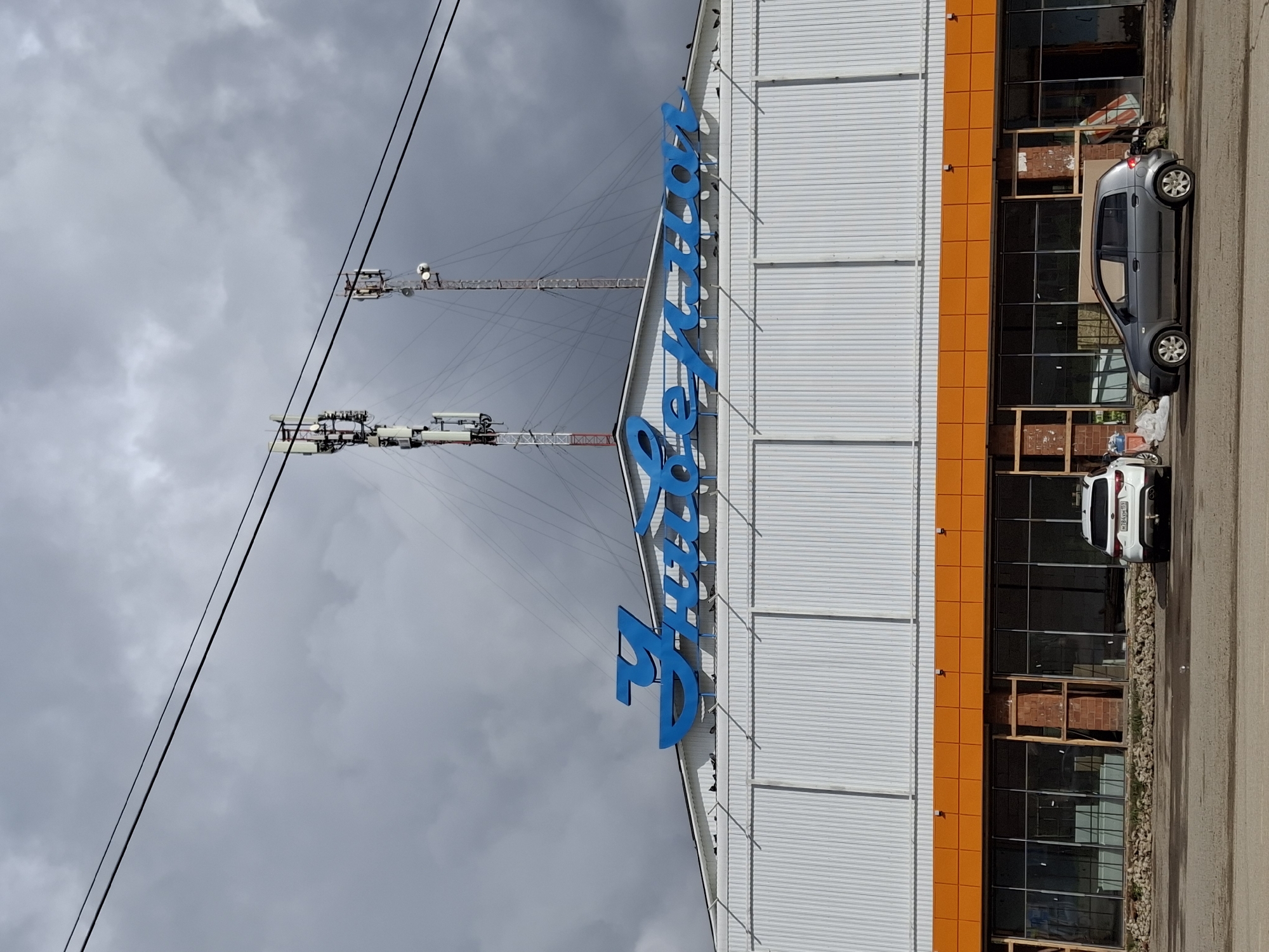 This image captures a view of a commercial building with a large blue sign in Russian Cyrillic script spelling "ГУБЕРНСКИЙ" across its facade, which translates to "Gubernskiy" or "Provincial." The building appears to be a store or market, characterized by its white corrugated metal exterior and a vibrant orange band running above a row of large, dark windows. Some of these window frames are under construction or have exposed brickwork. In the foreground, a gray hatchback car is parked on a paved area, and to its right, a white SUV is visible. Overhead, two cell towers rise against a heavily clouded, gray sky. The location is identified as Elovo, Russia.