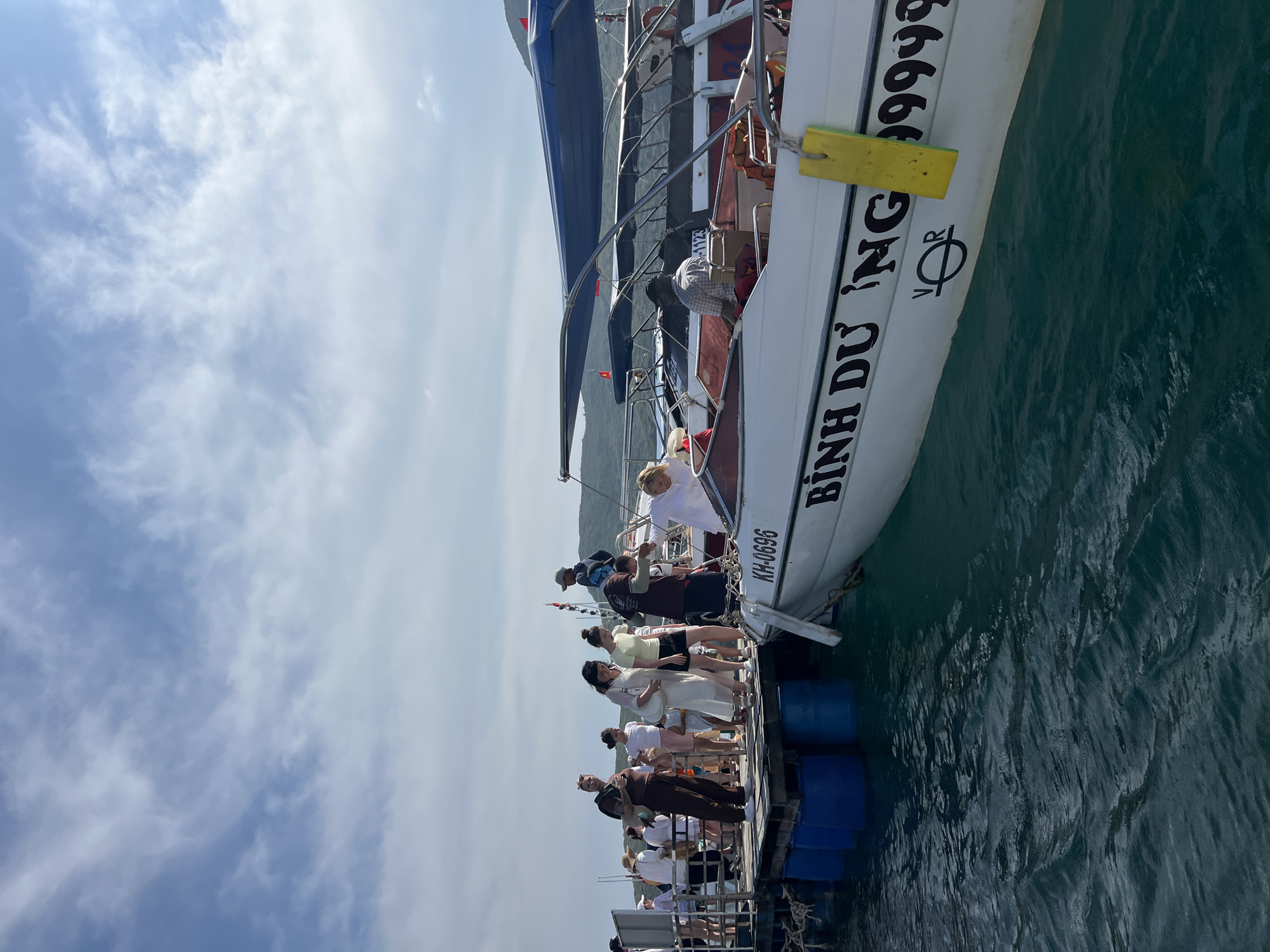 A horizontal, long shot depicts a tour group of approximately 20 people walking towards a white boat named "Binh Du." The boat is tied to a dock made of wooden planks and blue plastic barrels. A few people are already on board. The boat is white with a dark blue awning and has a yellow fender. On the side of the boat, it reads "Binh Du Ng" with the letters "VOR" below it, followed by the number "9999." The license number "KH-0696" is visible at the stern. The location is Nha Trang Ward, Vietnam, as the boat name suggests. The sky is partly cloudy.