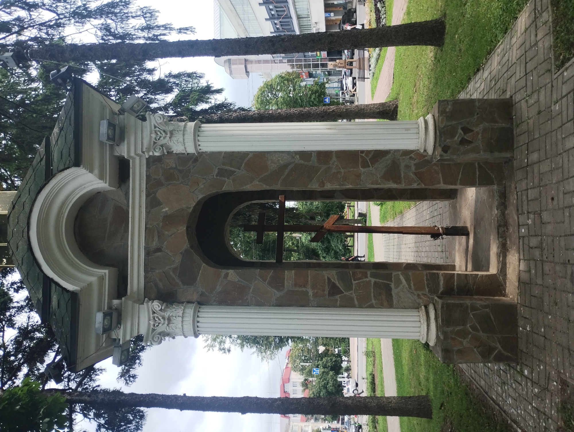 A stone-faced, arched alcove with two white columns topped with ornate capitals stands on a paved walkway. Within the alcove, a wooden cross is positioned vertically. The alcove is surrounded by green grass and trees, and a paved street with cars and buildings is visible in the background. The scene appears to be in a public park or square. This is likely the Chapel of the Holy Cross located on Voskresenskaya Street in Sumy, Ukraine.