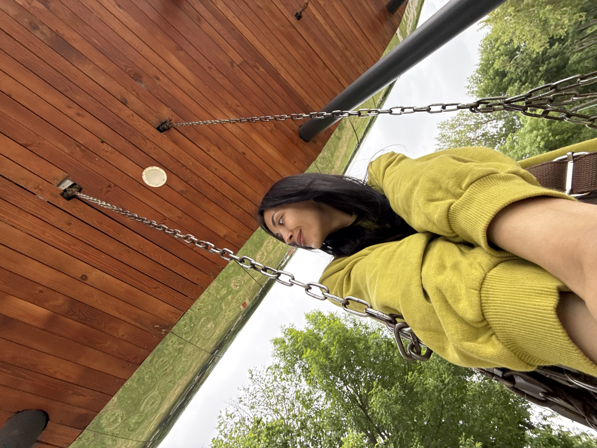 The image captures an outdoor scene from a low, upward-looking perspective, rotated approximately 90 degrees clockwise. A young woman with long, dark hair is the primary subject, visible on the right side of the frame. She is seen from her upper torso, wearing a vibrant lime green long-sleeved sweatshirt, and looks upward and to the left with a gentle, relaxed smile. A brown strap with a silver buckle, part of a swing or suspended seat, is visible near her arm.
Dominating the upper left portion is a sturdy wooden canopy or ceiling, constructed from reddish-brown planks. Several sets of metal chains descend from the wooden structure, anchoring into the wood and extending towards the lower right, indicating a swing mechanism. A circular, white recessed light fixture is embedded into one of the wooden planks. A thick, dark grey metal support bar runs across the mid-upper section of the frame.
Beneath the wooden structure and alongside the chains, a highly reflective, distorted surface captures the surrounding green foliage, creating a fluid, rippled pattern. Beyond these structures, an overcast sky is visible, along with the leafy canopies of green deciduous trees. The scene depicts a daytime moment of leisure in what appears to be a contemporary park or recreational area in Votkinsk, Russia. No visible text is present.