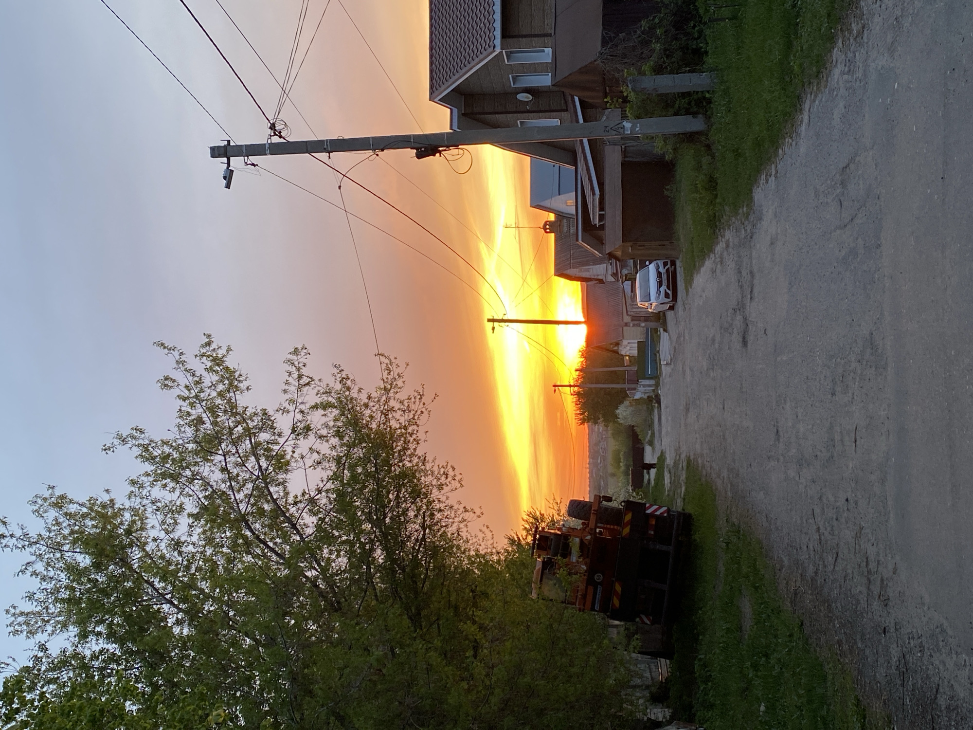 The image captures a street scene during sunset in Ulyanovsk, Russia. A dirt road runs from the bottom right to the middle ground, flanked by green grass and vegetation on either side. A large tree with green leaves is visible on the left side of the road. A parked, dark-colored truck is located along the roadside on the right, behind the tree. Overhead power lines stretch across the sky from left to right. The setting sun casts a bright, orange glow that illuminates the sky and the buildings lining the road. Houses are visible along the road, with a white car parked in front of one. The light of the setting sun is especially bright in the distance down the road.