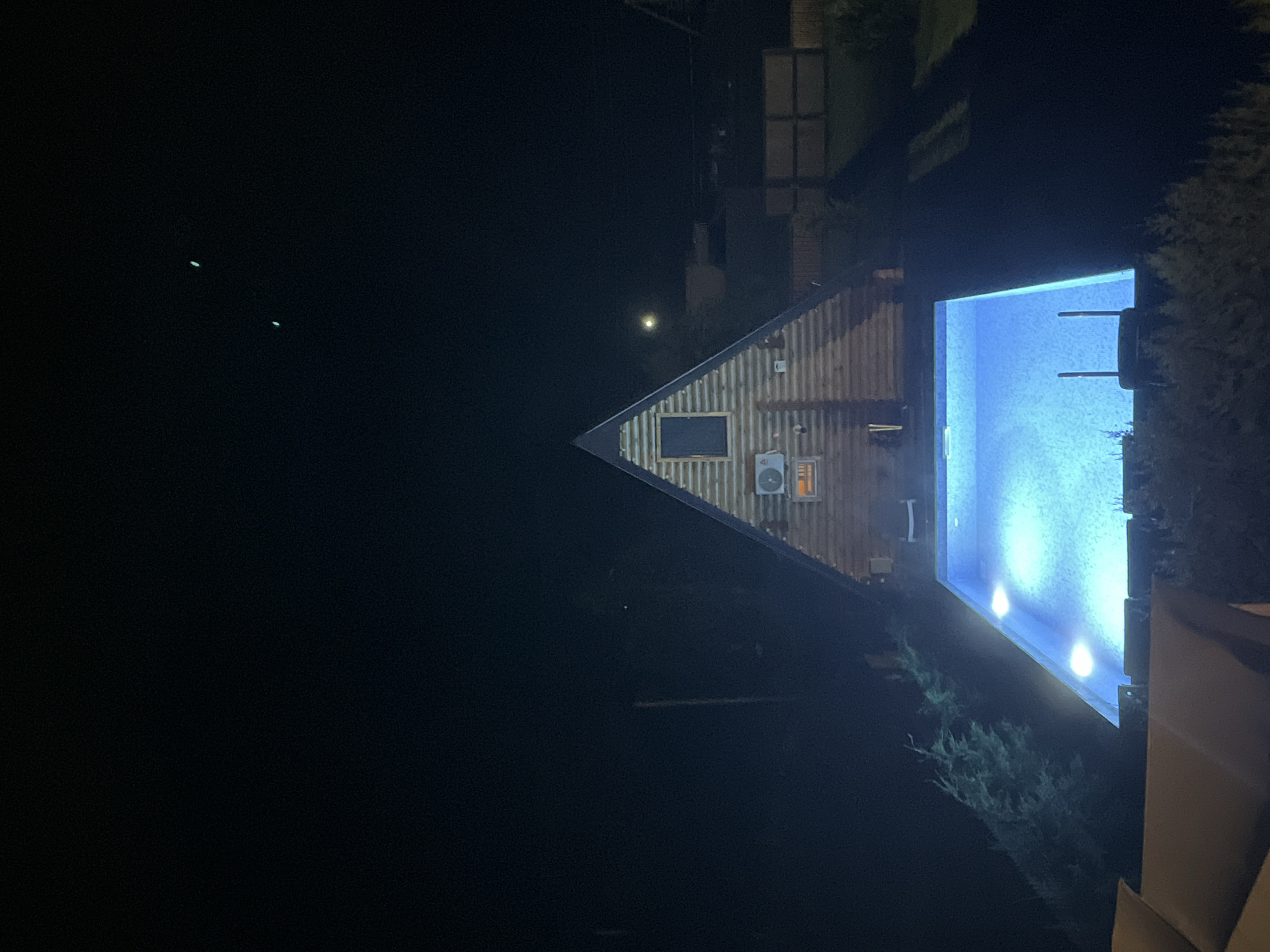 This is a nighttime, eye-level, medium shot of a triangular wooden cabin with a glowing blue swimming pool in front of it. The cabin has a dark door and window at the peak of its roof, and an air conditioning unit mounted on the exterior. The swimming pool is illuminated by underwater lights and appears to be empty of people. A few lounge chairs are visible in the foreground. The scene is set at night in a dark, outdoor environment. There are no people visible in the image. The specific location in the city of Tba, Georgia, is not discernible from the image.