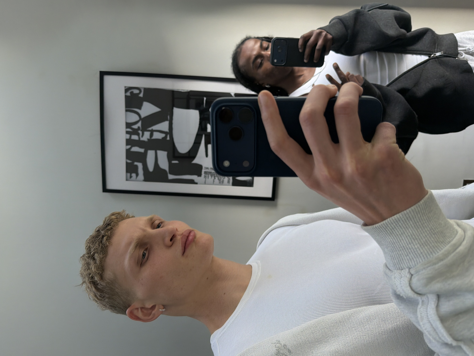 Two young men are taking selfies in what appears to be a modern apartment. The man in the foreground, on the left, has short blonde curly hair and is wearing a white long-sleeved shirt. He is holding up a dark blue smartphone to take a selfie. In the background, reflected in a mirror, is another young man with dark hair and a white tank top, wearing a dark grey zip-up hoodie. He is also holding up a smartphone, taking a selfie. Behind the man in the foreground, a framed abstract artwork hangs on the wall. The overall environment suggests an urban, likely residential setting. There is no indication of this being in London, United Kingdom.