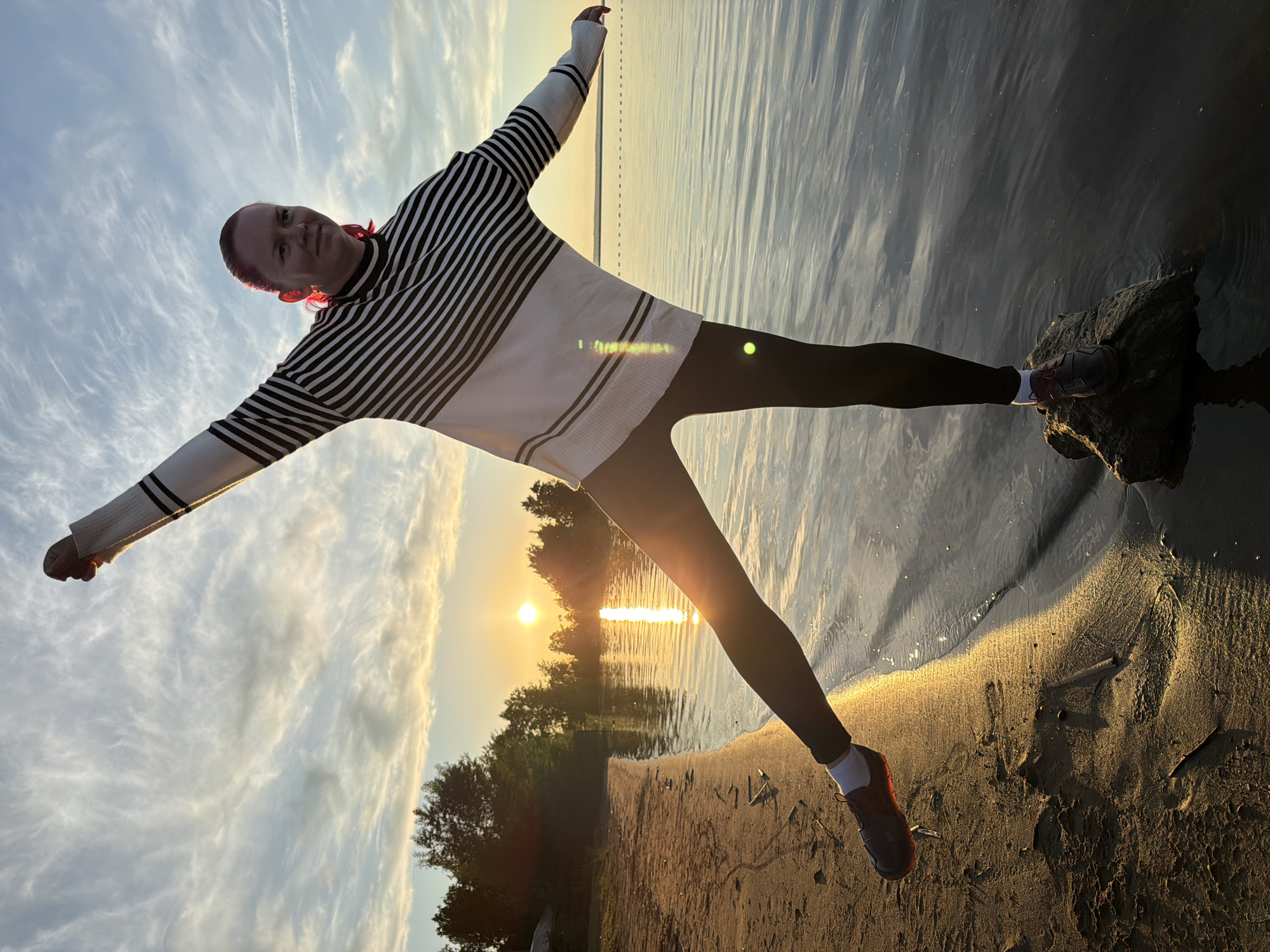 This image depicts a person striking a balanced pose by a tranquil body of water at sunset in Chaikovsky, Russia. The central figure is an individual with short, possibly reddish-pink hair, dressed in a white long-sleeved sweater with black horizontal stripes, dark leggings, and sneakers. Their arms are outstretched horizontally, creating a "T" shape, as if embracing the vastness of the scene or maintaining balance. The person looks towards the viewer with a subtle smile, conveying a sense of serenity and connection with nature.
The setting is a picturesque natural landscape, likely the Kama River or a local reservoir, characterized by calm waters reflecting the vibrant colors of the sky. The person's right foot is planted on a dark, mossy rock partially submerged in the water, while their left foot rests on the sandy-muddy shore. The sun, a bright golden orb, is positioned low on the horizon, casting a brilliant, shimmering path of light across the water and bathing the shoreline in a warm, golden glow. A dark treeline is visible in the distance across the water, silhouetted against the bright sky.
The sky above features scattered, wispy clouds in soft blue and orange hues, indicative of either a sunrise or, more likely, a sunset, given the intense warm light. The overall mood is peaceful and expansive, capturing a moment of quiet enjoyment in a beautiful natural environment. No visible text is present in the image.
