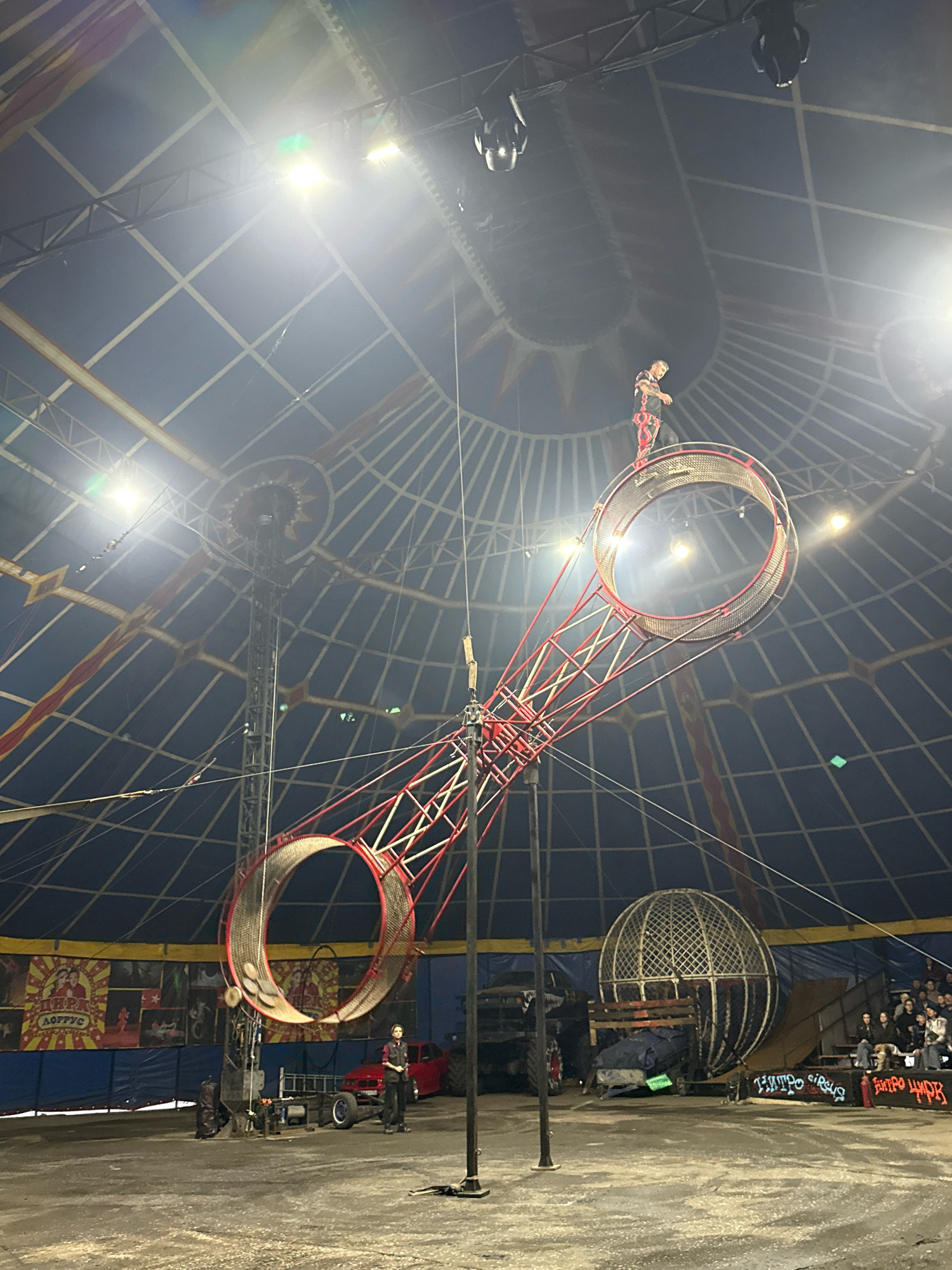 The image depicts a circus tent in Rusanovo, Russia, illuminated by spotlights from above. A performer in a red and black costume stands atop a large, circular metal structure, which is part of a larger, elongated device with two connected rings. This apparatus is suspended in the air by cables. In the background, a large metal sphere, commonly known as a "globe of death," is visible, along with a red vehicle and spectators seated on bleachers. The ground appears to be dirt or sand. The overall scene suggests an ongoing circus performance.
