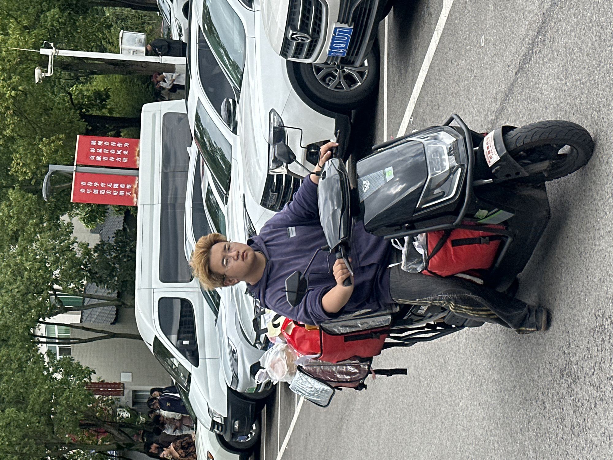 A young adult male sits on a black electric scooter in what appears to be a parking area or street in Jiangjin, China. He has short, light blonde hair and wears glasses, a blue hooded sweatshirt, and dark pants. His expression is neutral, as he looks slightly upwards and to his left. The scooter is equipped with multiple red and silver insulated bags and clear plastic bags, characteristic of a food or package delivery vehicle.
In the background, several white vans or SUVs are parked. One car prominently displays a blue and white license plate with the characters "A-01077," suggesting a local Chongqing vehicle (A-prefix often indicates Chongqing). Lush green trees and a building are visible behind the vehicles. A tall pole with surveillance cameras and two vertical red banners featuring white Chinese text stands next to a white van.
The text on the banners reads:
"展现青春作为 彰显青春风采 贡献青春力量" (Show youth's actions, display youth's demeanor, contribute youth's strength)
"青春孕育无限希望 青年创造美好明天" (Youth fosters endless hope, youth creates a beautiful tomorrow)
Another smaller, partially obscured sticker on the scooter's front fender reads "绿源" (LüYuan), likely the brand of the electric scooter. The setting appears to be daytime, under an overcast sky or diffused light, as there are no harsh shadows. The individual seems to be taking a break or waiting for an order.