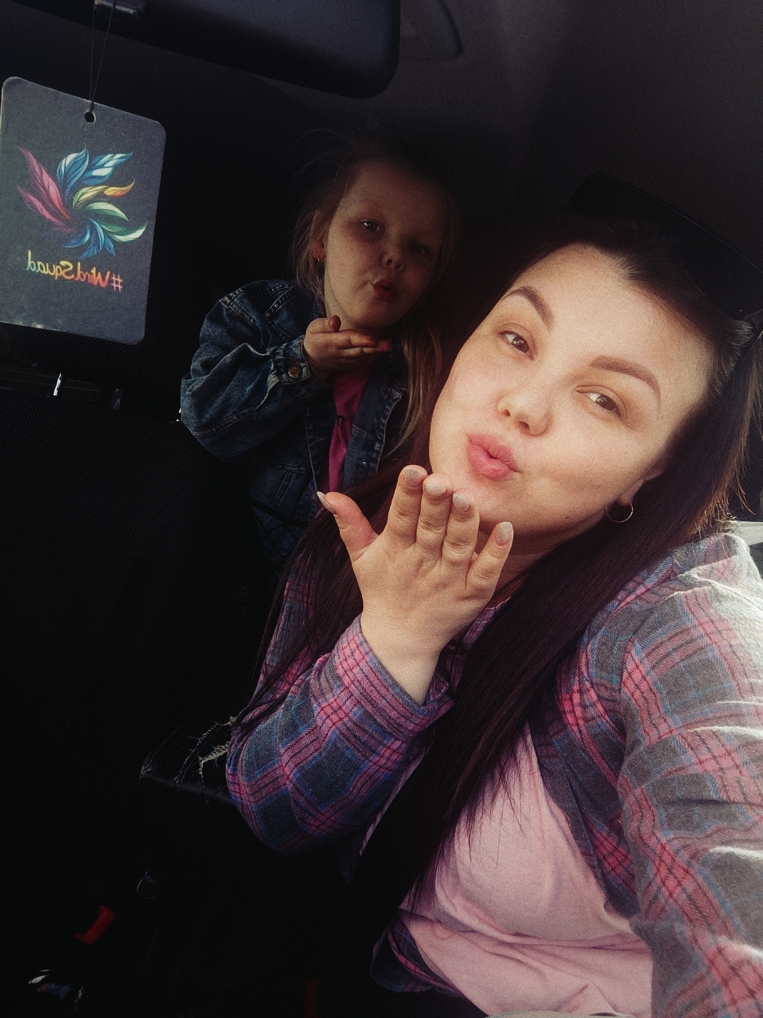 A selfie taken inside a vehicle in what appears to be Chaikovsky, Russia, features two females. In the foreground, an adult female with brown hair and a plaid shirt puckers her lips and holds her hand up as if blowing a kiss. A younger female, likely a child, wearing a denim jacket, mirrors the same action in the background. A car air freshener hangs from the rearview mirror, partially obscuring the child. The interior is dark, with black seats and parts of the car's interior visible. The subjects seem to be engaged in a playful or affectionate interaction.