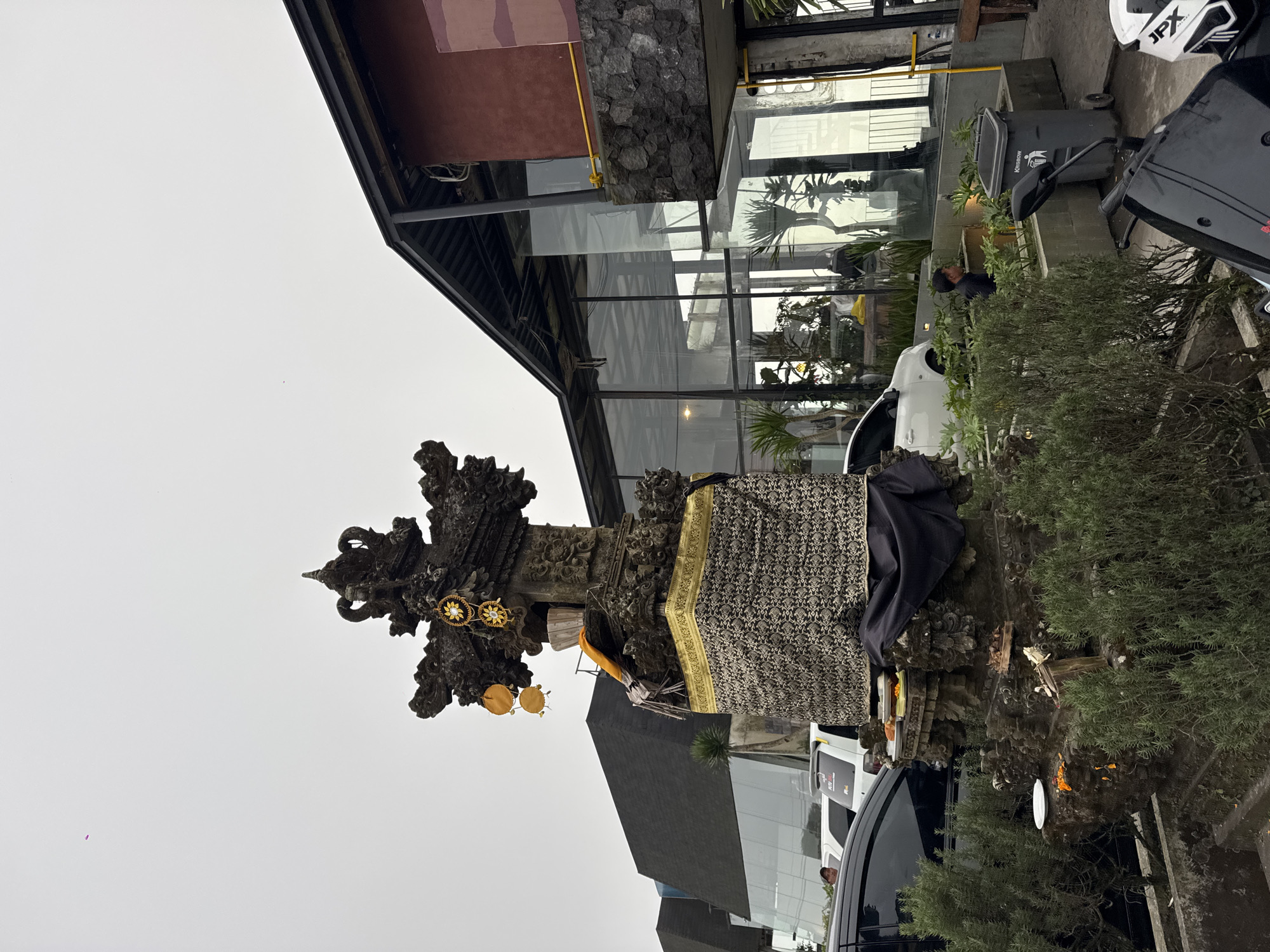 In Kintamani, Indonesia, a Balinese stone altar is positioned in front of a modern building with large glass windows. The altar is adorned with offerings, including plates of food and yellow flowers, and is draped with a black cloth. A gold and black patterned fabric wraps around the base of the altar. Behind the altar, the building features a dark, angled roof and red walls. Several vehicles are parked in the vicinity, including black and white cars and a scooter. Lush green bushes surround the altar. The sky is overcast, and the overall atmosphere appears calm and observant.