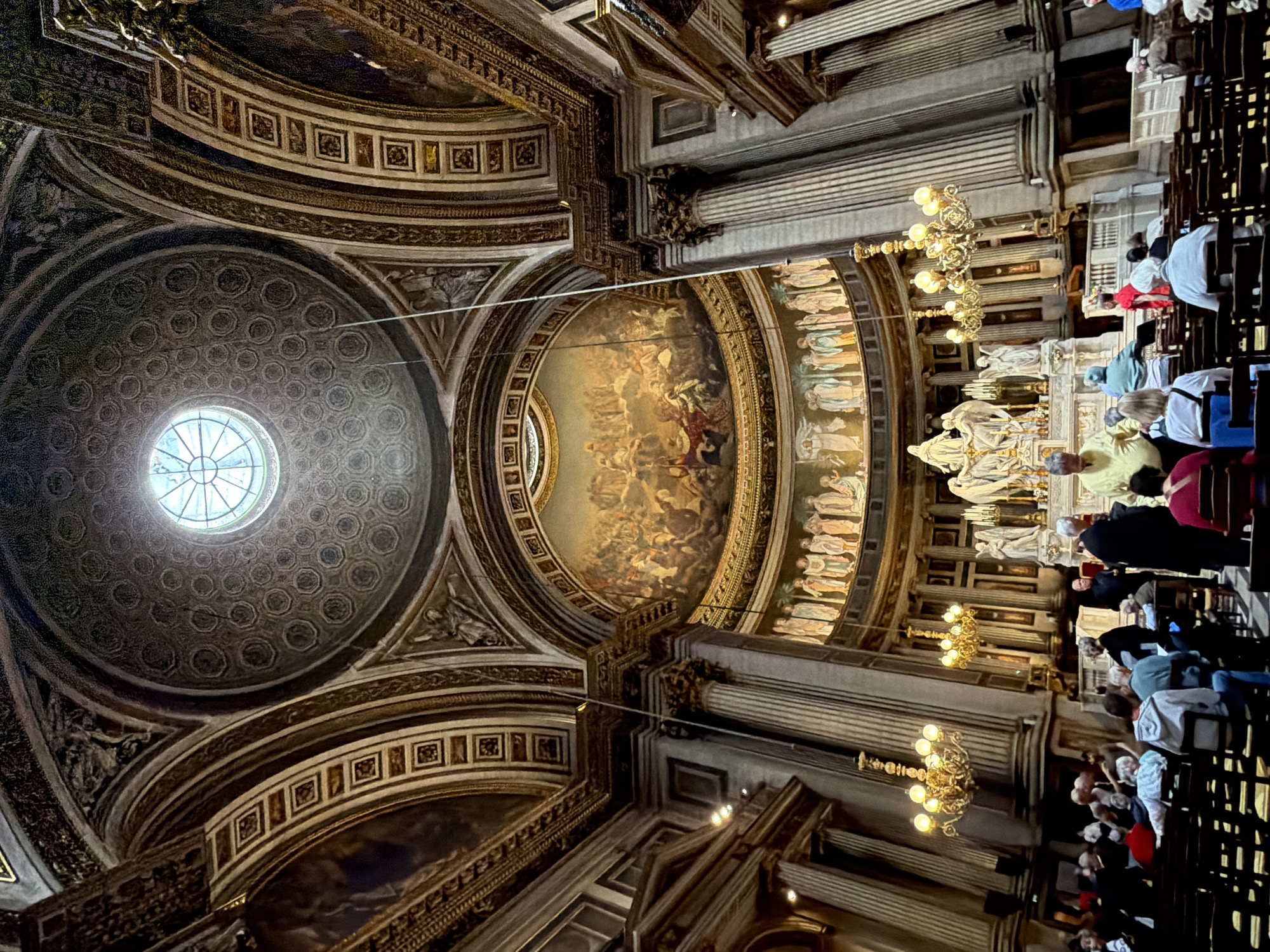 The image depicts the interior of the Madeleine Church in Paris, France. The vast, ornate dome of the church is visible, featuring a circular window at its apex and intricate patterned decoration. Large, detailed paintings adorn the curved walls and ceiling, depicting religious scenes. Several grand columns with Corinthian capitals line the nave, supporting the arched structure. Golden chandeliers with multiple lights hang from the ceiling. Below, a congregation of people is seated in rows of dark wooden chairs, facing the altar. Some individuals stand and move about the church. The altar area is visible in the background, featuring statues and religious iconography. The overall atmosphere is one of solemn grandeur and historical significance.