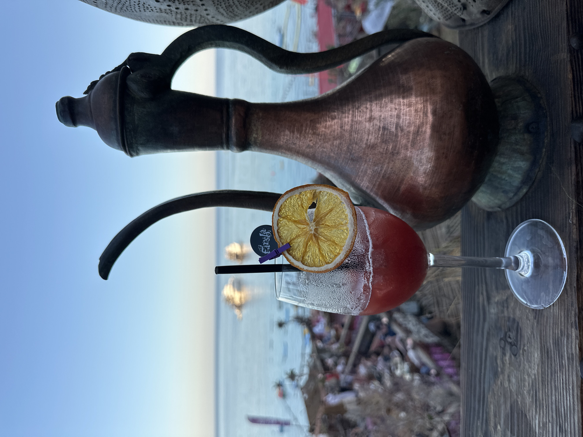 A refreshing red cocktail in a stemmed glass, garnished with a dried orange slice and a black straw, rests on a rustic wooden surface. A small black tag, secured by a purple mini clothespin to the orange slice, displays the word "Farsha" in white stylized text. To the left of the glass stands a tall, ornate copper pitcher with a slender spout and handle, exhibiting a traditional, weathered appearance.
The scene is set outdoors, overlooking a calm, light blue sea that extends to a distant horizon under a clear, bright sky. The background reveals a hazy shoreline or beach area with indistinct figures or structures, suggesting a leisure or resort environment. The lighting suggests late afternoon or early evening, contributing to a relaxed and tranquil atmosphere. The cocktail glass shows condensation, indicating a cold beverage. The overall ambiance and the traditional copper pitcher hint at a unique cultural setting, consistent with the provided location context of Sharm El Sheikh, Egypt.