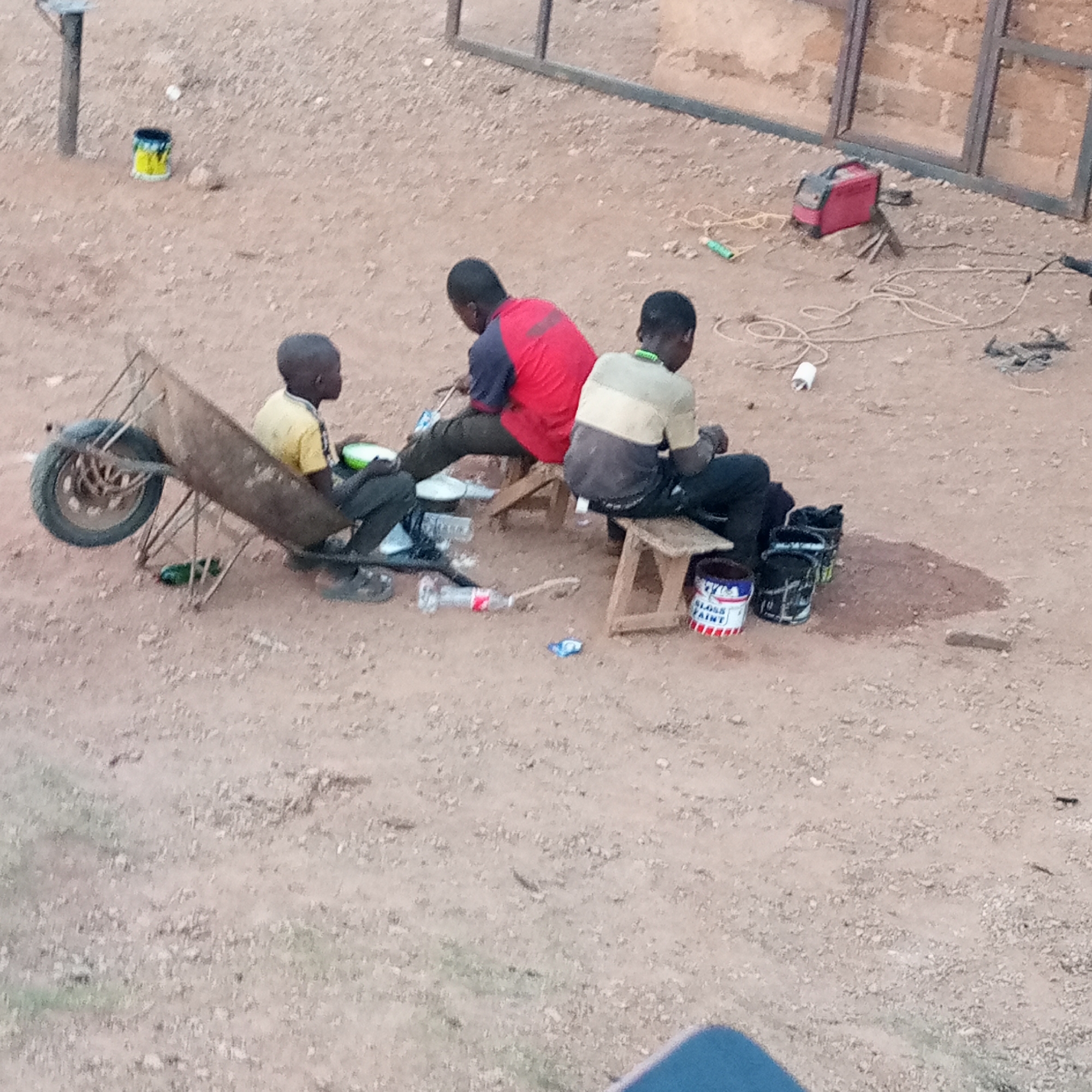 Three young men are sitting outside on small wooden stools in what appears to be a dusty or gravel-covered area in Kuru Dungus, Nigeria. The man on the left is sitting in a wheelbarrow. Next to him is an open bottle of soda and some plates. A man in a red shirt sits on the second stool. The man on the right is sitting on a stool with four paint cans sitting on the ground next to him. There is a welding machine and metal frames behind them. A yellow paint can is in the distance.