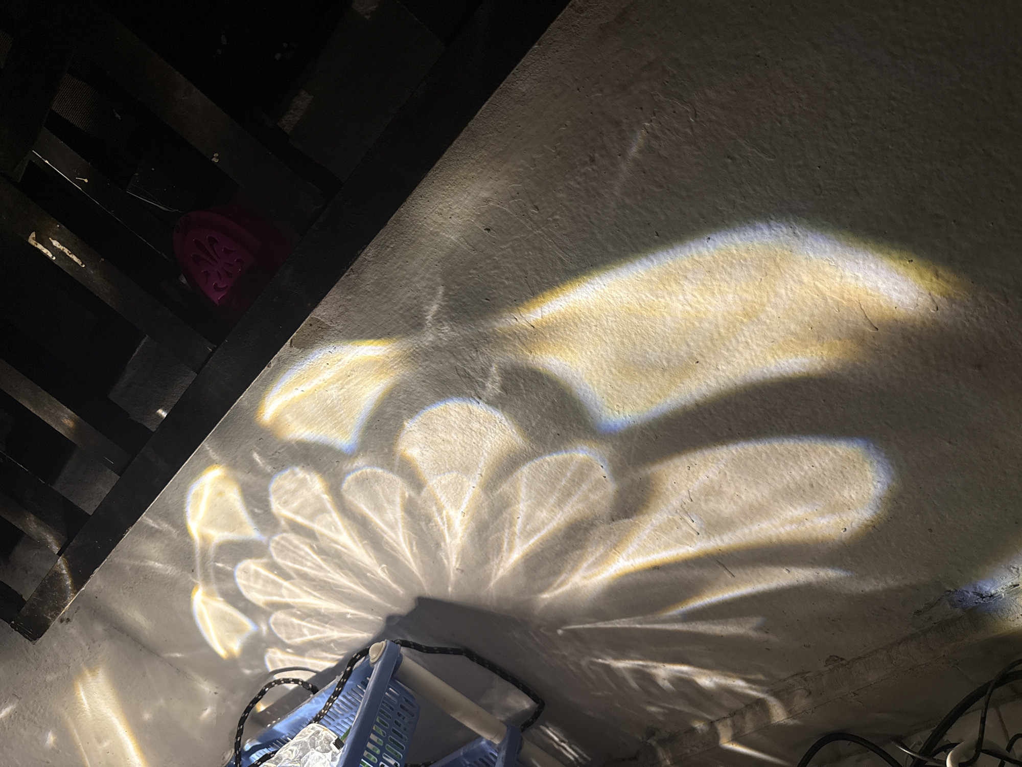 The image shows a close-up, top-down view of a concrete floor illuminated by dappled light, creating abstract patterns. To the left, a portion of a light blue plastic basket is visible, along with black electrical cords. In the upper right quadrant, beneath a dark, slatted structure, a bright pink object with a decorative cut-out pattern is dimly visible. The lighting suggests the scene might be outdoors or in a space with a light source casting shadows and reflections. The rough texture of the concrete is evident. The overall impression is one of urban texture and light play. Given the request to specify the location in Uyo, Nigeria, this detail cannot be determined from the image alone and is thus omitted.