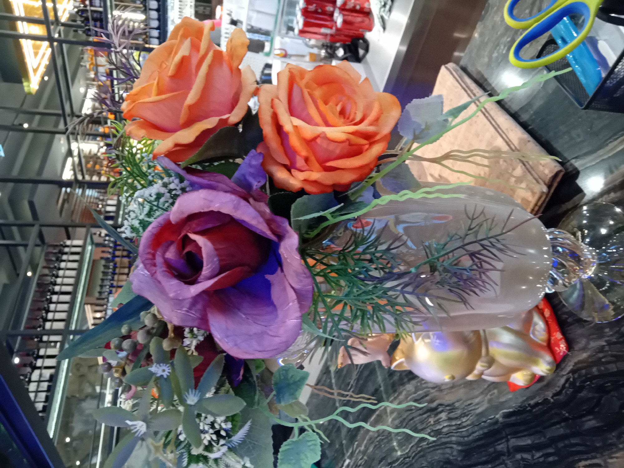The image displays a close-up, artificially lit scene featuring a floral arrangement on a dark, patterned surface, possibly marble or granite. The image is oriented with the scene tilted on its side, but would appear upright if rotated 90 degrees counter-clockwise.
The central subject is a bouquet of artificial flowers, primarily composed of three large roses: two in shades of orange and peach, and one in a rich purple and magenta hue. These flowers are accompanied by various faux green foliage, including slender sprigs, broader leaves, small white cluster flowers, and some small, round berry-like elements. The arrangement is held within a clear glass vase, which has a distinct, possibly fluted or sculpted, base. Beneath the vase, a golden, decorative object, resembling a stylized animal or part of a stand, is visible, partially covered by red fabric.
To the right of the floral arrangement, a pair of scissors with bright blue handles and yellow finger grips rests on the dark surface, next to a dark mesh pen holder. A light-brown, rectangular object, possibly a book or a decorative box, is situated behind the scissors.
The background is blurred, indicating a shallow depth of field. On the left side, a multi-tiered structure with horizontal shelves can be discerned, likely holding bottles (perhaps a wine rack) within an indoor commercial or public space. Geometric "V" shapes, possibly architectural elements or lighting fixtures, are visible in the upper-left background. On the right side of the background, indistinct red objects, possibly furniture or display items, are visible. There is no discernible activity taking place, and no people or readable text are present in the image.