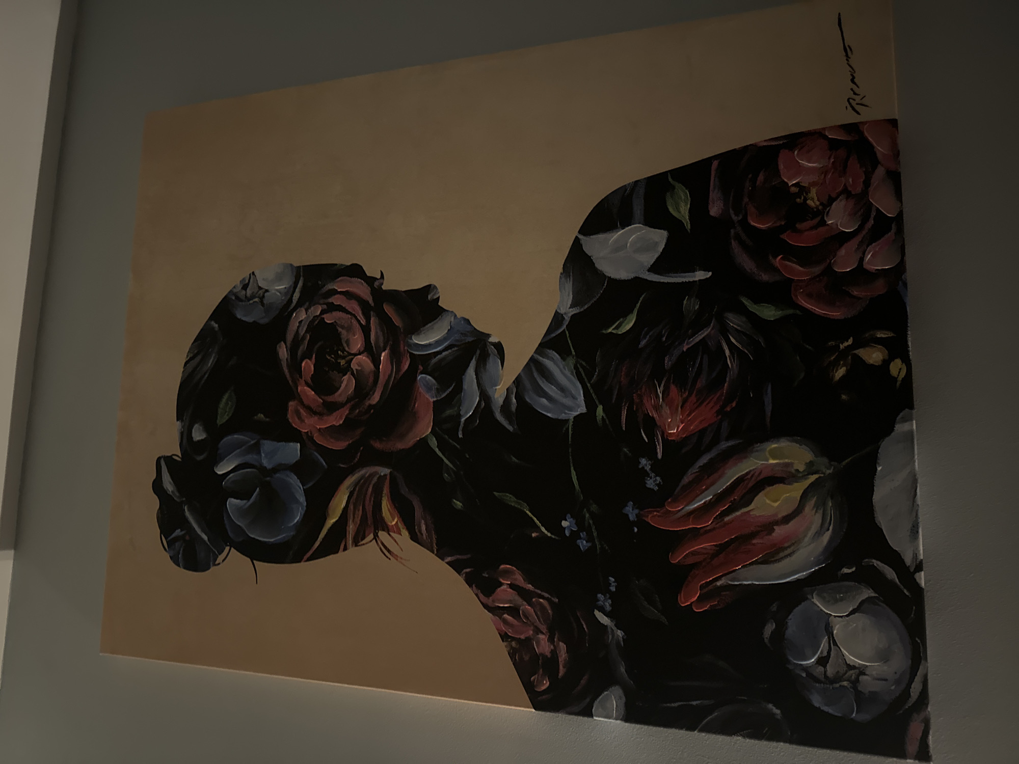 The image shows a painting hanging on a light blue wall. The painting depicts a silhouette of a woman's head and upper body filled with various flowers in shades of red, blue, and white against a beige background. The painting appears to be signed in the upper right corner. The location is not specified.