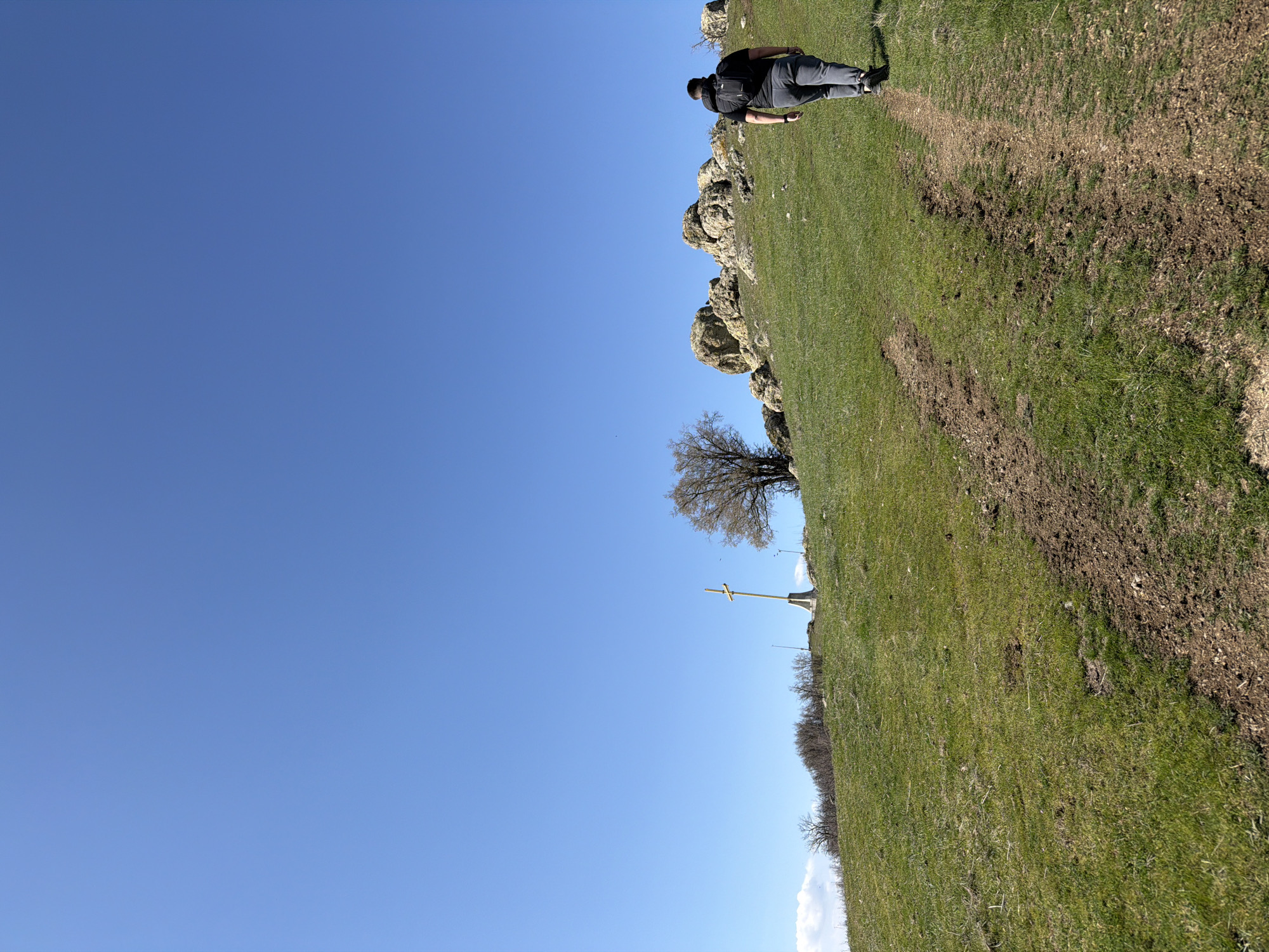 A landscape photo shows a clear blue sky occupying most of the image. A grassy hill occupies the rest of the image, with a path or trail visible on the right side. A person is walking along the trail at the top right of the frame. A rock formation is visible at the top of the hill. A metal cross is situated in the middle of the hill. The image is likely taken in Bata, Bulgaria, based on the presence of the metal cross.