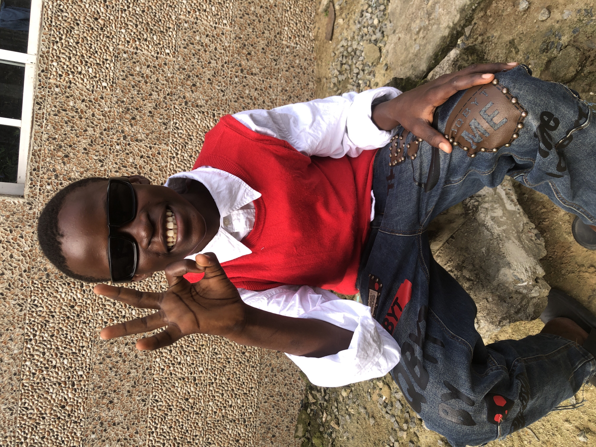 A young Black male, wearing sunglasses, a white collared shirt, a red vest, and distressed denim jeans, sits outdoors on rocks, giving a "peace" or "okay" hand gesture. The background is a textured wall and a window frame. The scene appears to be in Ado, Nigeria, based on the context provided.