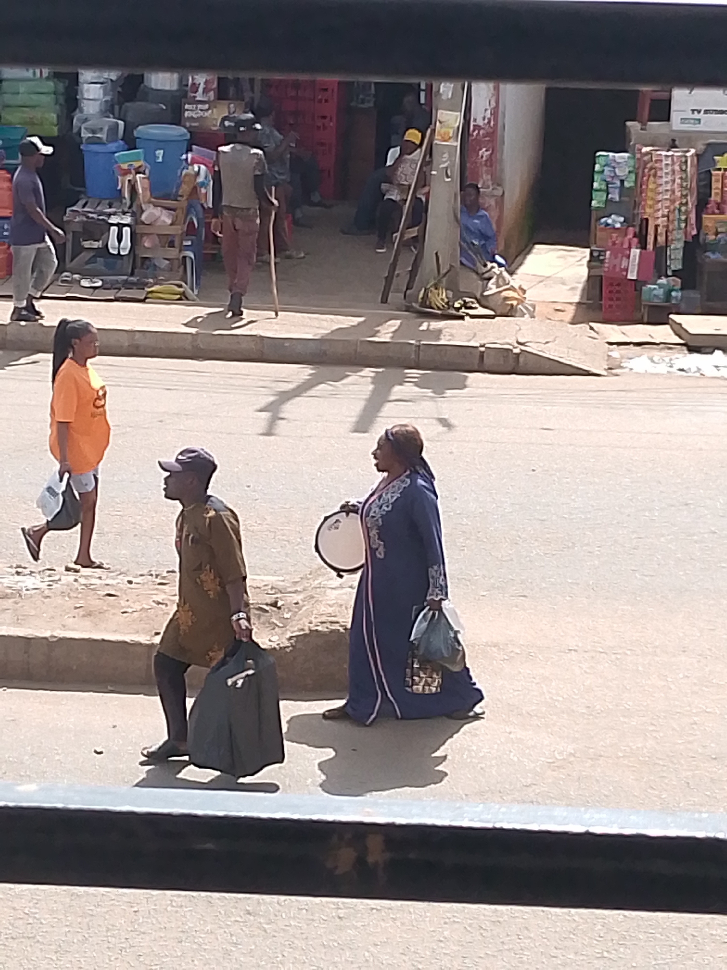 The image shows a street scene in Bukuru, Nigeria, with several people and a market stall in the background. In the foreground, a woman in an orange t-shirt and shorts, with her hair in a ponytail, walks to the left, carrying a plastic bag. Next to her, a man in a brown patterned tunic and dark pants, wearing a baseball cap, walks to the right, carrying a black bag. Further to the right, a woman in a blue patterned robe walks to the right, carrying a plastic bag and a small drum. In the background, a man with a cane walks to the left, and other people are visible near market stalls. The stalls display various goods, including crates of drinks, plastic containers, and other merchandise. The scene appears to be a busy street, likely during daytime, with the sun casting shadows. The overall impression is a snapshot of daily life in a Nigerian town.