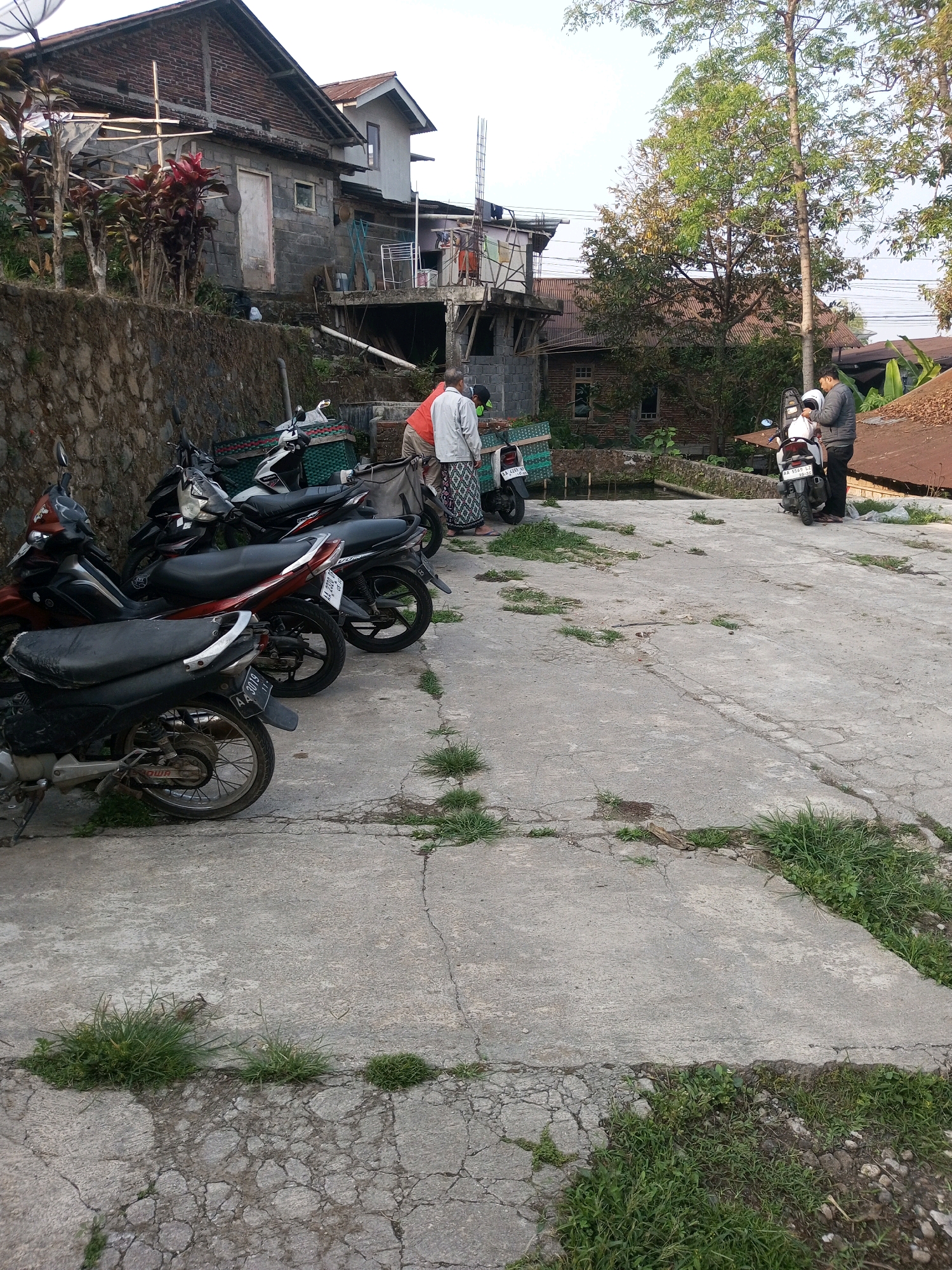 The image depicts a parking area in Kalibeber, Indonesia, with several motorcycles parked in a row. In the background, there are buildings, including a two-story structure under construction, and trees. Two men are standing near a white motorcycle, one holding a helmet. Another man, wearing a white shirt and patterned trousers, is standing near a green crate. The ground is made of concrete with cracks and patches of grass growing through it. The lighting suggests it is daytime, with sunlight illuminating the scene.