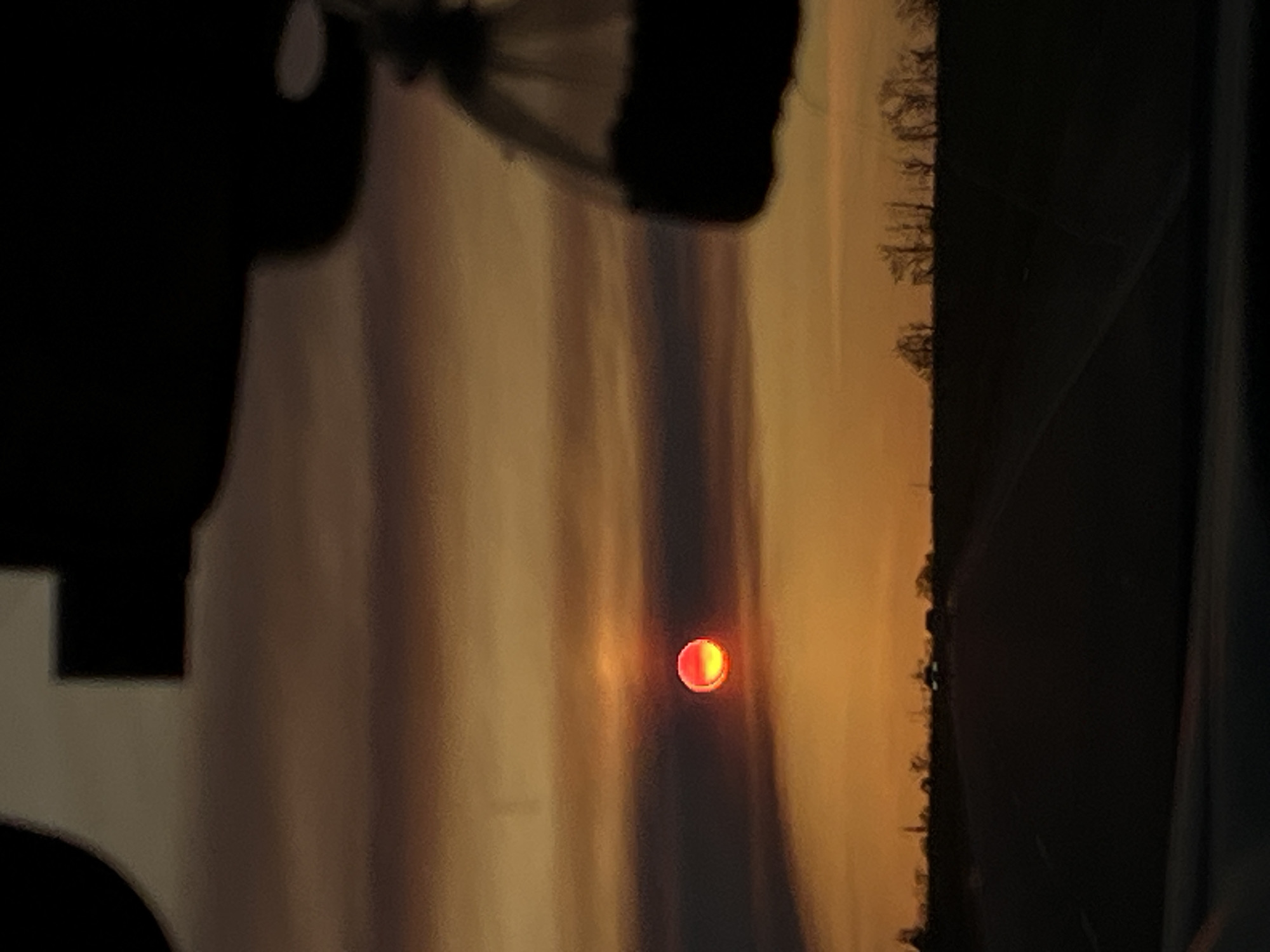 The image displays a vibrant sunset viewed through a foreground obstruction. The sky is filled with warm hues of orange, red, and yellow, transitioning into darker, muted tones higher up. Streaky clouds are visible, adding texture to the colorful expanse. A distinct, bright red-orange disc, identified as the sun, is prominent in the middle of the frame, appearing partially obscured or low on the horizon.
Below the illuminated sky, a thin, jagged line of dark silhouettes represents a distant treeline, indicating the horizon. The scene is partially framed by large, dark, out-of-focus shapes on the left and top sides of the image (when correctly oriented). These shapes suggest the photograph was taken from behind an object, possibly a window frame, a structural element of a building, or part of a vehicle.
The activity taking place is a sunset. The time of day is clearly evening. No specific weather conditions beyond atmospheric haze contributing to the sunset colors are discernible. There are no visible location cues within the image itself to suggest Vyselki, Russia, and no discernible text. The image captures a natural phenomenon viewed from a confined or partially obscured perspective.