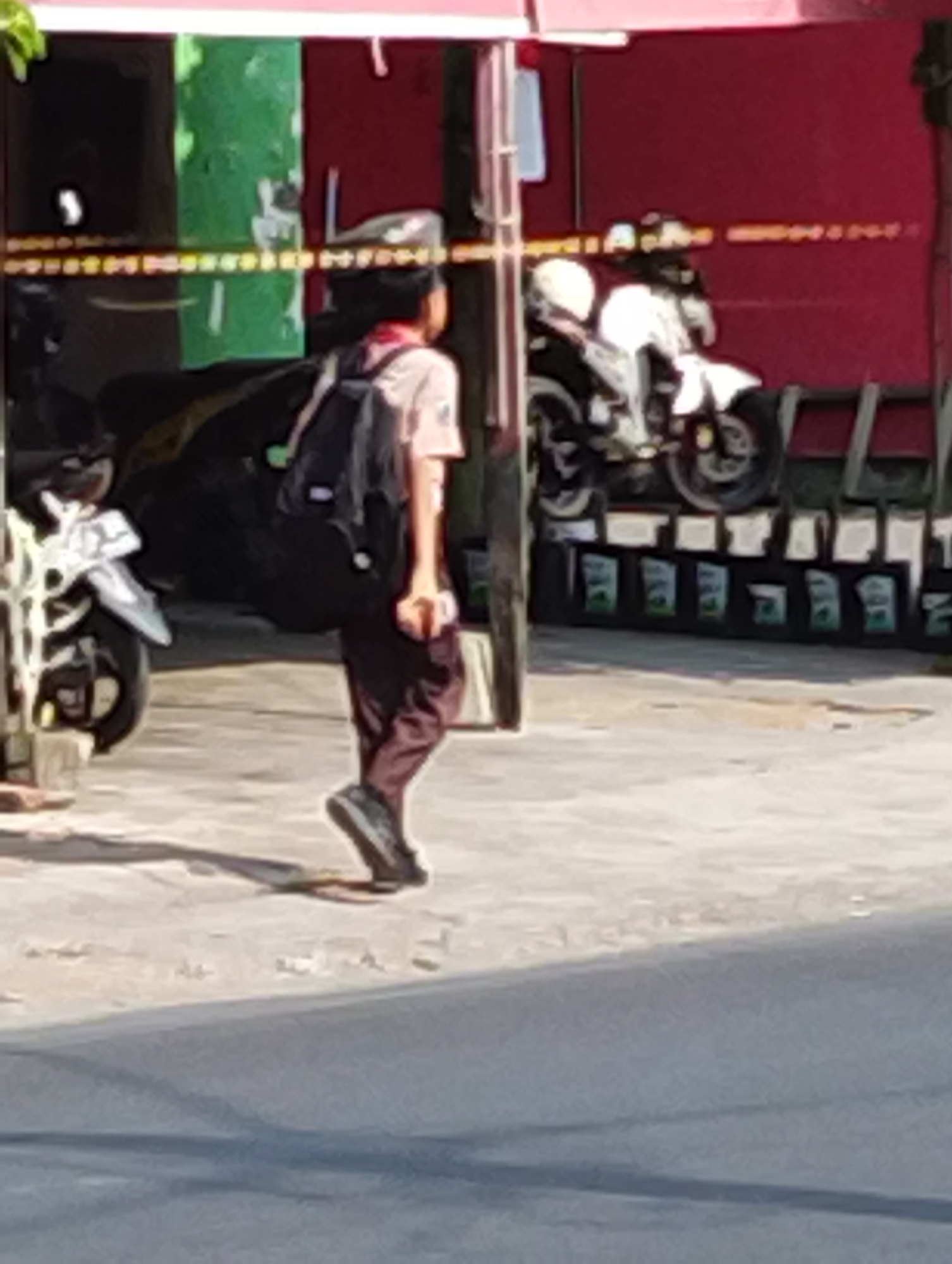 The image depicts an outdoor scene in Manaruwi, Indonesia, featuring a young person wearing a light-colored shirt, dark pants, a backpack, and a cap, walking from left to right across a paved area. To the left, a motorcycle is partially visible, with its front wheel and fender showing. In the background, there is a red structure, possibly a building, with a green section on the left. A white motorcycle with a rider in a helmet is parked in front of the red structure. Several dark objects, possibly crates or boxes, are arranged in a row in front of the red structure. The ground is a light-colored pavement, with shadows cast by the subjects and objects. The overall impression is a snapshot of a street scene in a localized area.