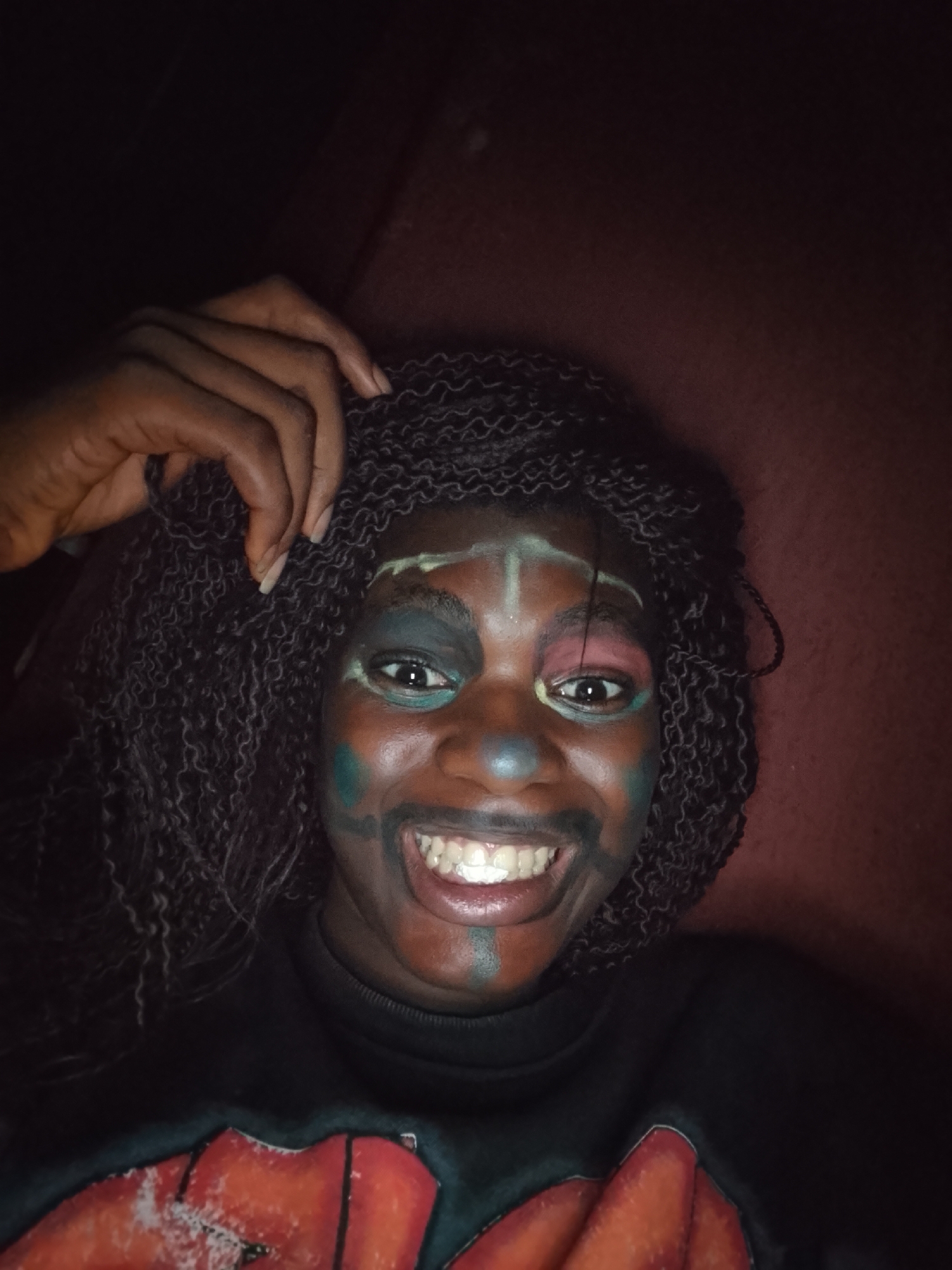 A portrait-style image captures a person with dark skin, likely female, smiling broadly directly at the camera. Her dark, braided hair frames her face. She is adorned with colorful face paint, featuring light green/white on her forehead and down her nose, dark green/teal around her left eye and on her left cheek, pink/red eyeshadow around her right eye, and a blue/grey dot on her nose. Black lines create an exaggerated, wide smile extending from the corners of her mouth, showcasing her white teeth. Her right hand, with neatly trimmed fingernails, rests near her temple, touching her hair. She is wearing a dark, long-sleeved top, possibly a sweater, with a large, vibrant red and orange design on the chest. The background is dark and out of focus, revealing hints of a reddish-brown surface, suggesting an indoor setting with low lighting, likely taken at night. The subject appears happy and cheerful, posing for the photo.