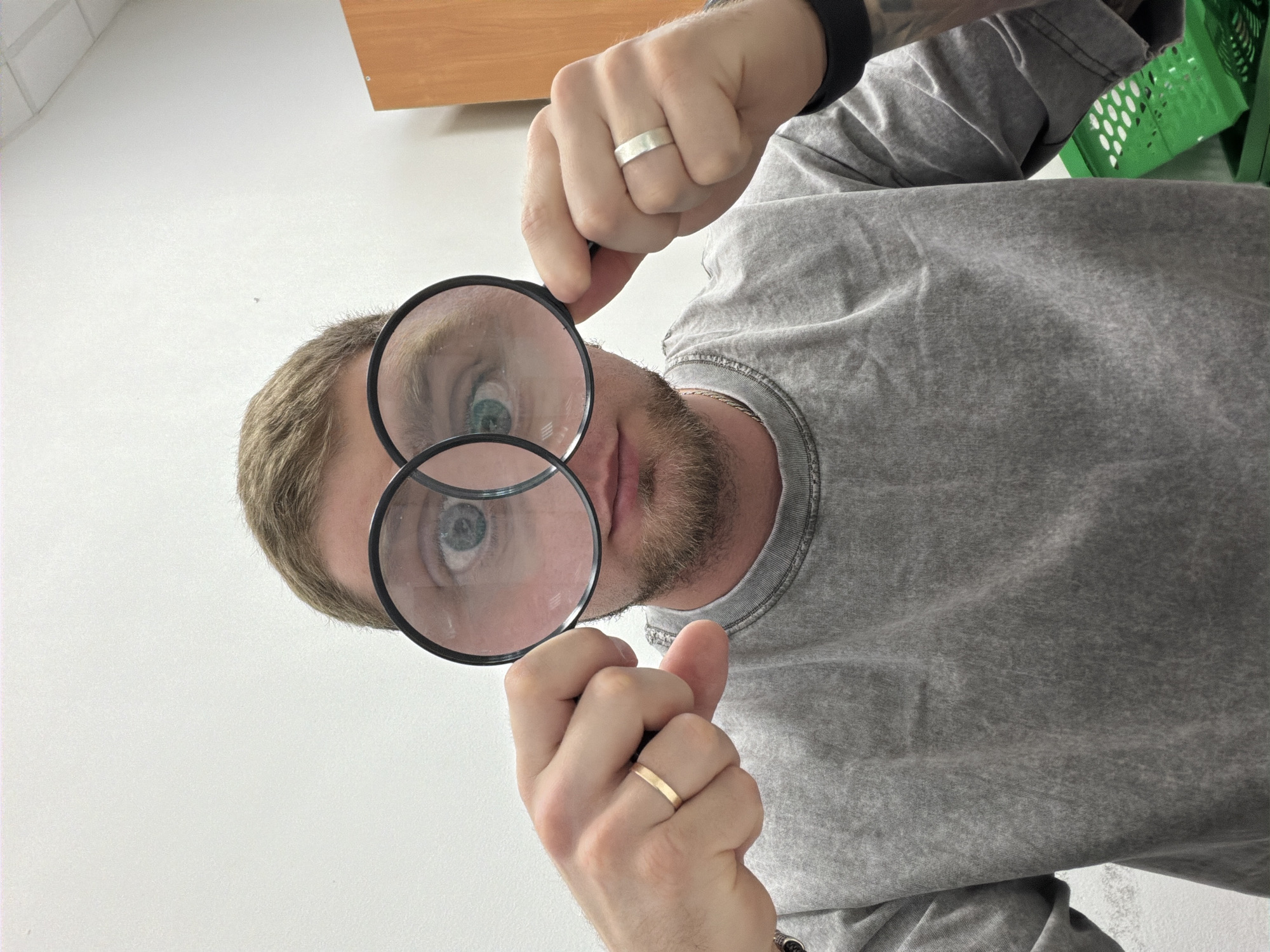 A person with short blonde hair, a beard, and light eyes is holding two magnifying glasses in front of their eyes. The person is wearing a grey t-shirt, a black smartwatch, and a ring on each hand (one silver, one gold). The background includes a white floor, a wooden cabinet, and a green plastic basket. The person's face is angled upwards, as if looking directly at the viewer. The location is not identifiable.
