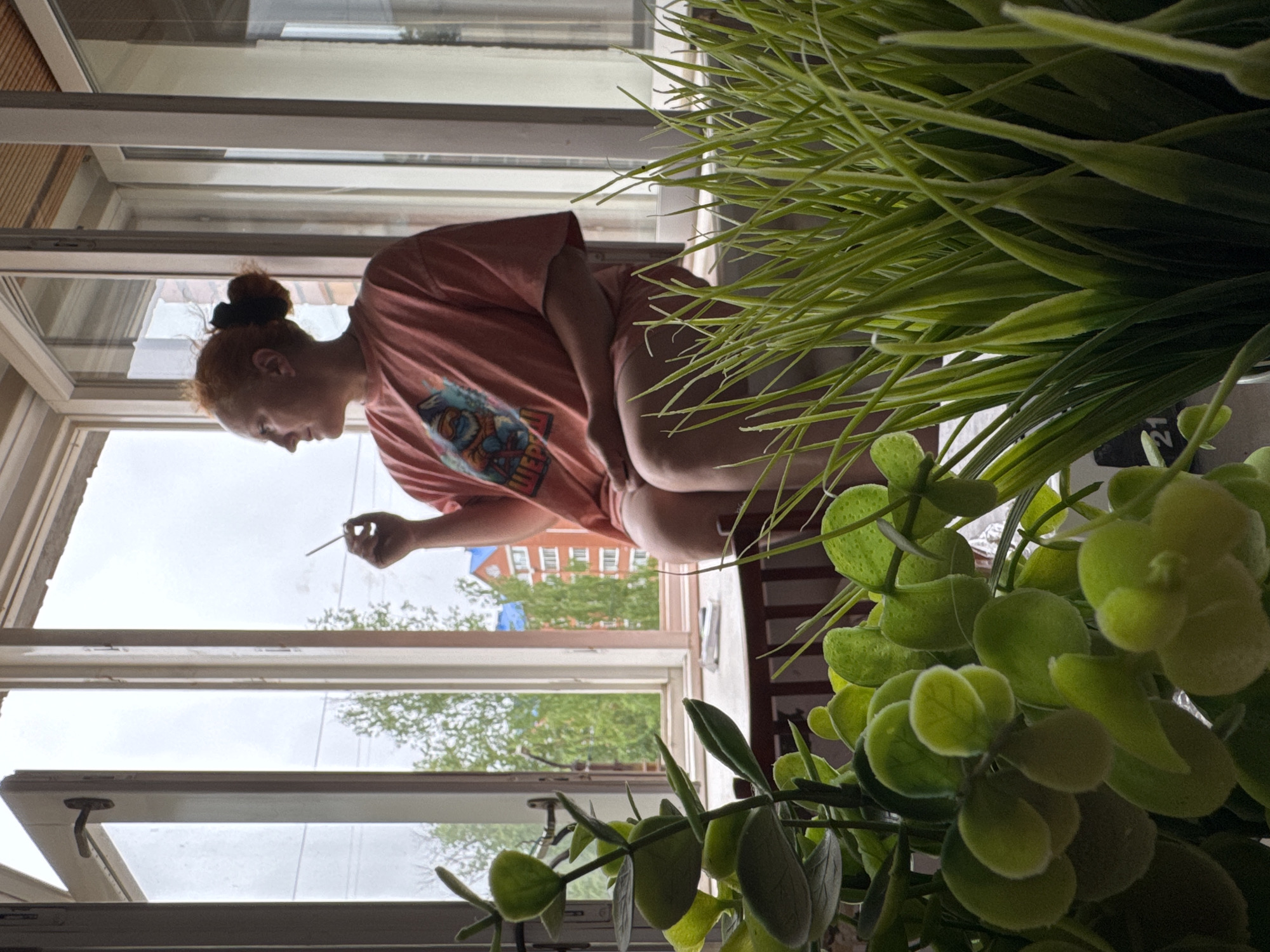 A woman with red hair pulled into a bun is sitting on a windowsill inside an apartment, smoking a cigarette. She is wearing a pink t-shirt. The window is open and overlooks a street with buildings and trees. In the foreground are two potted plants, with green foliage obscuring part of the scene. The location is Chaikovsky, Russia.