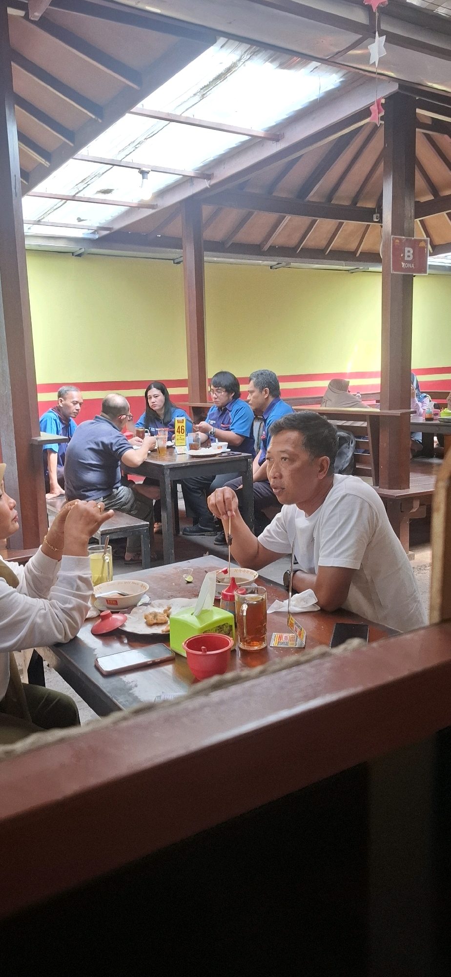 The image depicts an indoor dining area, possibly a restaurant or cafe, located in Colomadu, Indonesia. The setting appears to be covered by a wooden roof structure with translucent panels allowing natural light to filter through. Several tables with wooden chairs are arranged for patrons. In the foreground, a man in a white t-shirt is seated at a table, holding a skewer with food. A woman in a white long-sleeved shirt is also seated at the same table, gesturing with her hands. On the table, there are bowls, glasses of iced tea, a tissue box, a red container, and a smartphone. In the background, a group of people, mostly wearing blue shirts, are seated at another table, engaged in conversation and eating. One person is holding a table tent with the number "46". The walls of the establishment are painted yellow with a red stripe. A sign with "B ZONA" is visible on a pillar in the background. The overall atmosphere suggests a casual dining or gathering environment.