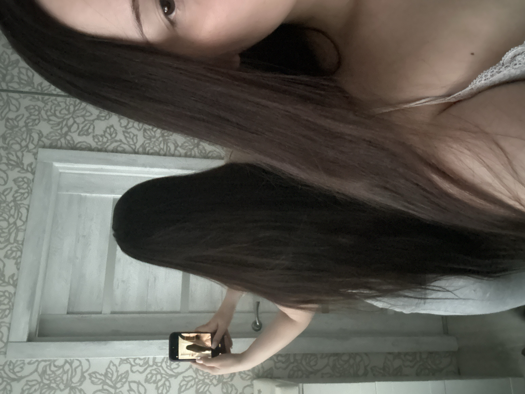 The image captures an indoor mirror selfie. On the right side of the frame, the subject's partial reflection reveals the right portion of her face, including a dark brown eye and eyebrow, along with her right shoulder and upper chest. She has long, straight, dark brown hair extending down her shoulder and is wearing a light-colored camisole with visible lace trim. A small mole is present on her upper chest.
On the left side of the frame, the subject's full reflection is visible from the back. Her long, straight, dark brown hair extends significantly down her back. She holds a dark-colored smartphone horizontally in both hands, positioned to capture her reflection in the mirror. The phone's screen displays a live camera feed reflecting her upper body and hair.
The background features a light-colored interior door with a silver-toned handle. The walls are adorned with light-toned wallpaper showcasing a floral or botanical pattern. The specific location in the city of Chaikovsky, Russia, cannot be visually confirmed from the provided image content.