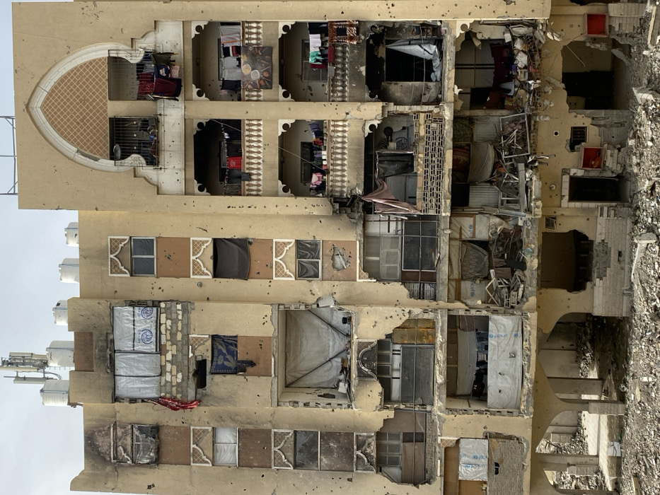 An image displays a severely damaged multi-story residential building in Khan Yunis, Palestinian Territory, exhibiting extensive structural degradation and numerous impact points. The light brown facade is pockmarked with bullet holes across all visible levels.
The building's roof features multiple white water storage tanks and at least one communication antenna. The upper floors show varying degrees of damage; some windows are shattered or boarded with improvised materials, including white sheeting, some bearing "UNICEF" logos. Burn marks are visible on the facade near the top left. Several balconies, characterized by ornate white balustrades, still have laundry hanging, indicating continued use or recent evacuation. A satellite dish is mounted on an upper-right balcony.
Mid-levels of the building feature large, irregular holes blown through the walls, exposing the interiors of multiple apartments. These exposed sections reveal rubble, debris, damaged furniture, and makeshift coverings. Lower levels of the building exhibit more catastrophic structural failure, with entire sections of walls collapsed, exposing shattered rooms and a significant accumulation of rubble and debris spilling onto the ground below. The ground level, originally supported by arches, is completely filled with debris. No people are visible in the scene.