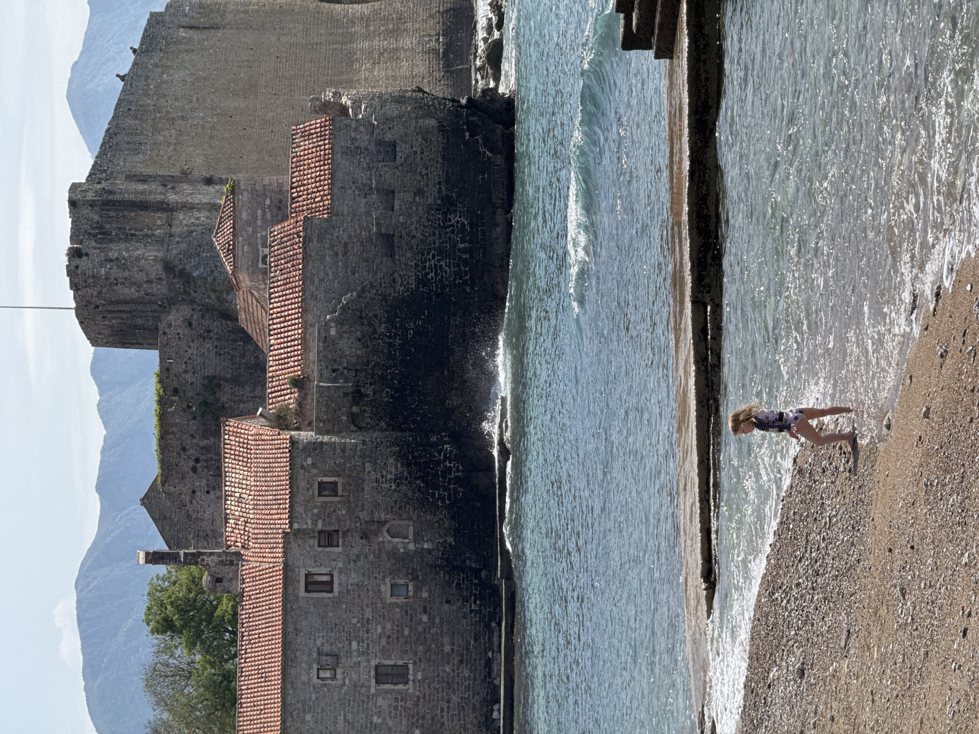 A sideways view depicts a sunlit beach beside a stone building complex in Budva, Montenegro. A young girl wearing a swimsuit stands in the shallow surf, where waves gently meet the pebble-covered beach. A concrete structure divides the beach and sea. Adjacent to the beach, aged stone buildings with red tile roofs are stacked against a larger stone wall structure. In the background, hazy mountains are visible. The sky is light blue.