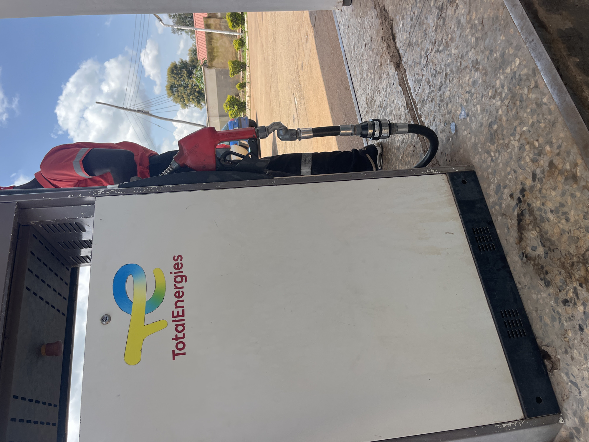 In Gura Top, Nigeria, a close-up shot captures a fuel pump at a TotalEnergies gas station. A person wearing a red shirt with reflective stripes, typical of station attendants, is partially visible. They are holding a red fuel nozzle connected to a black hose, which is positioned over the concrete ground. The side of the fuel pump is visible, displaying the TotalEnergies logo. The background shows a cloudy blue sky, a building with a red roof, and some greenery. The ground is made of textured concrete, with some dark stains and small patches of lighter liquid.