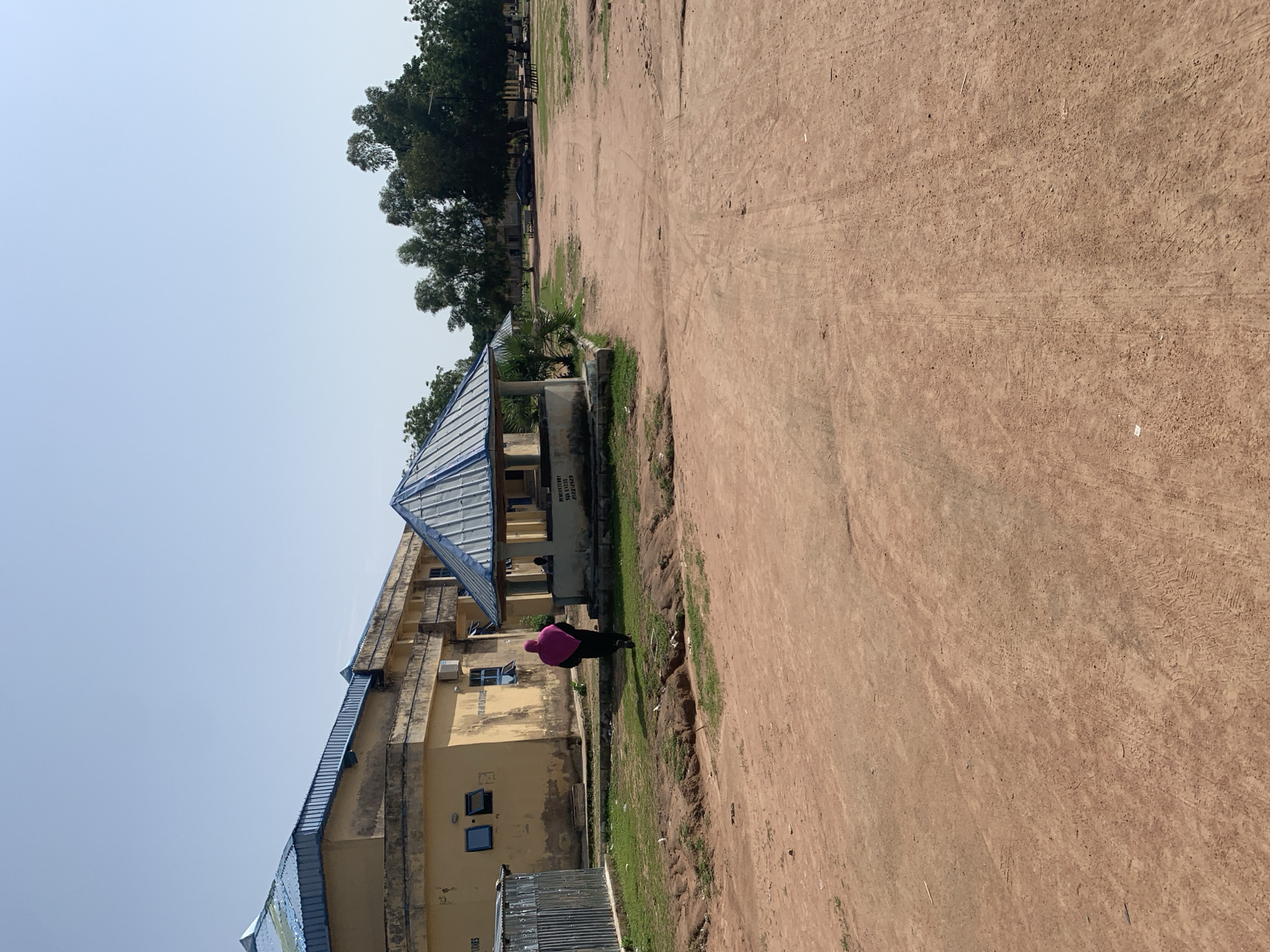 The image captures a side view of a light yellow, multi-story building with a blue metal roof, possibly a school or administrative building. A person wearing a pink headscarf and dark clothing is standing in a grassy area next to the building. The foreground consists of a wide expanse of reddish-brown dirt, showing tire tracks and debris, suggesting it's an outdoor area such as a courtyard or a sports field. In the background, there are trees and what appears to be more buildings. The sky is clear and bright, indicating a sunny day. A small, open-sided structure with a metal roof is visible near the building. Some text is visible on the building's wall, but it is partially obscured and illegible in this orientation. The scene appears to be set in Jalingo, Nigeria, under a bright sky.
