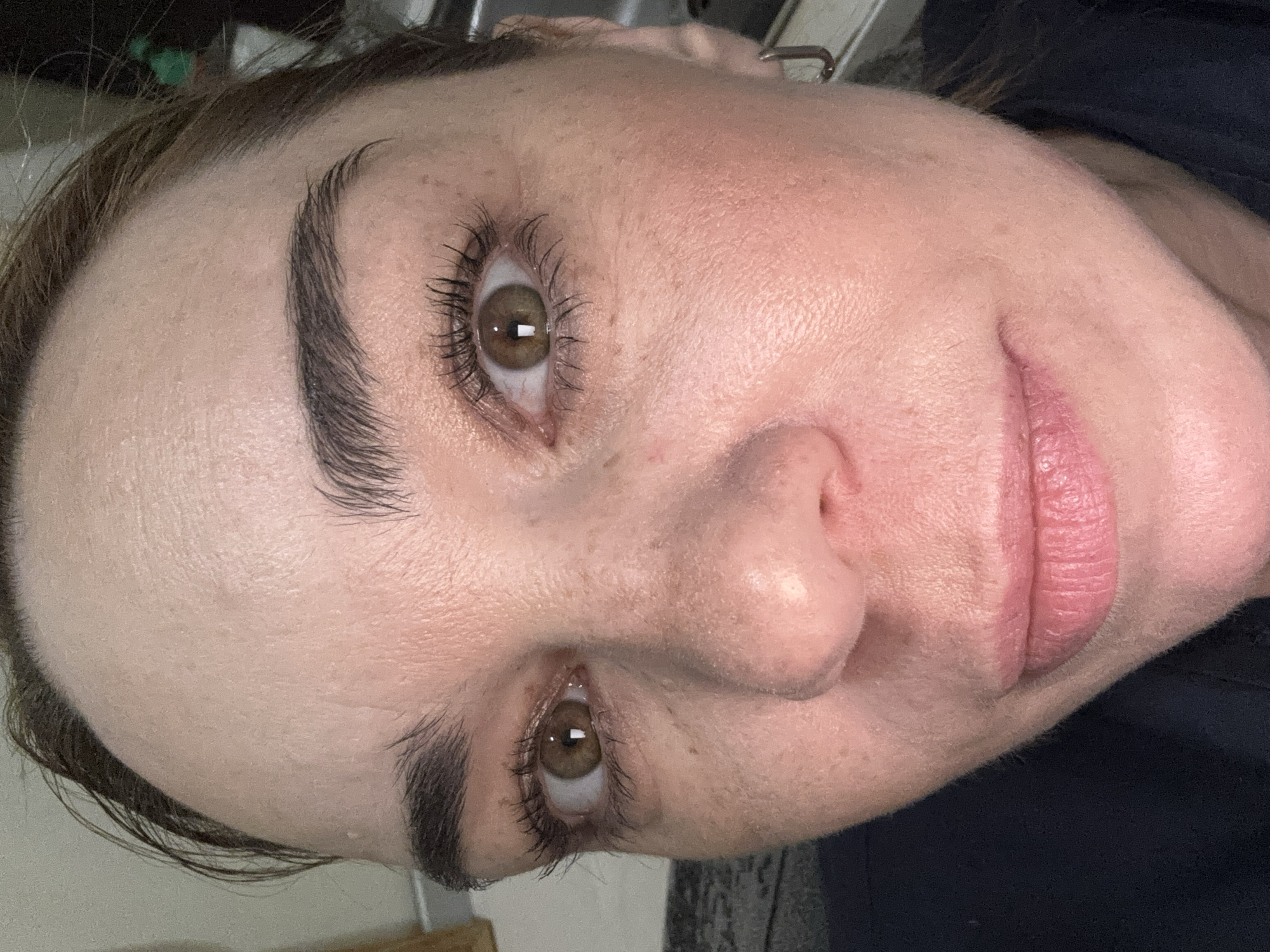 A close-up shot of a woman's face, tilted to the right. She has fair skin, brown eyes, and dark eyebrows. Her lips are closed and appear to be a light pink color. She appears to have light makeup on. The background is blurred and indistinct, with some dark fabric visible.