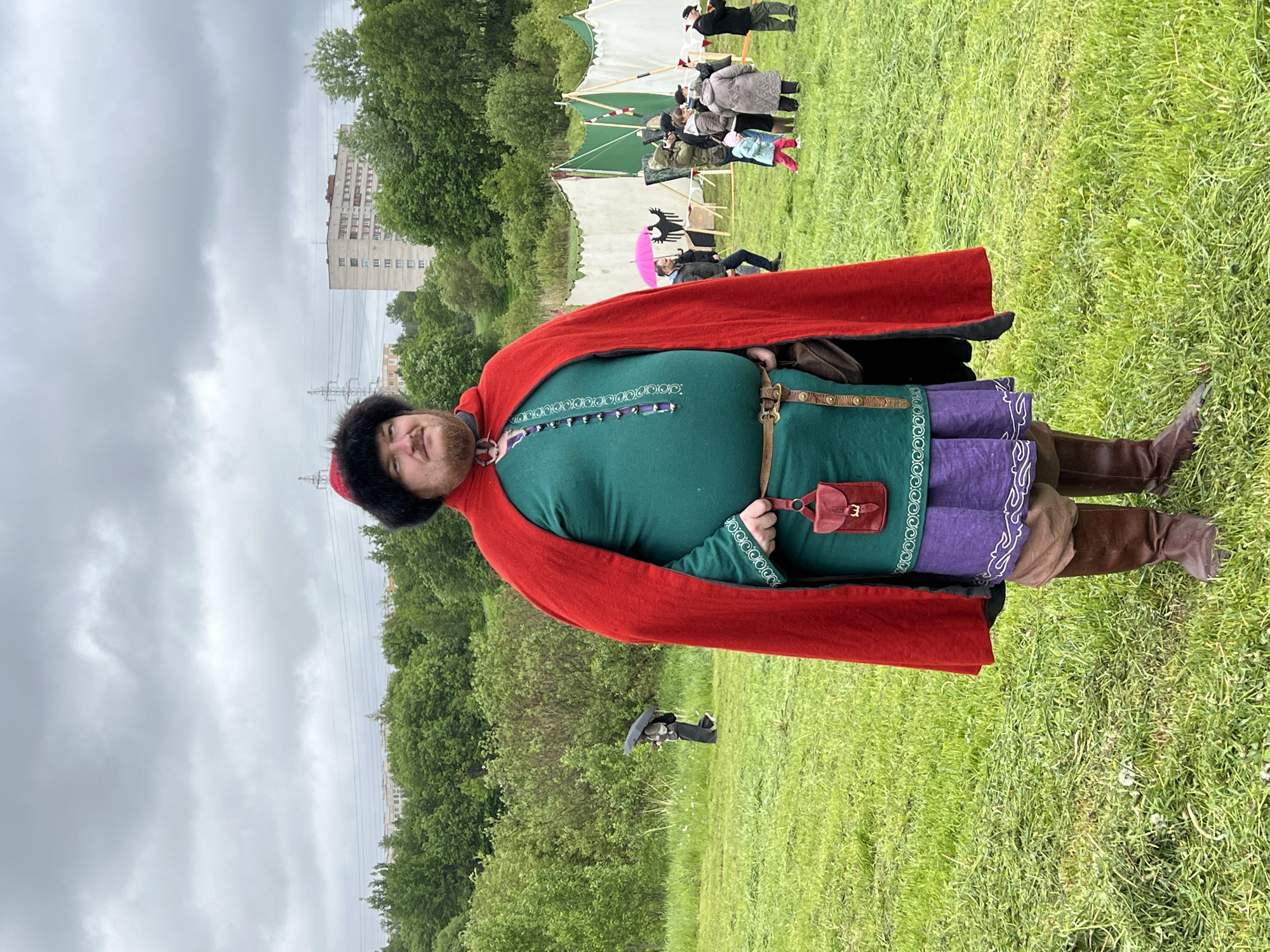A person is standing in a grassy field wearing a historical costume that includes a fur hat, red cape, green tunic, purple pants, and brown boots. The costume also features a small red pouch hanging from the belt. The setting is outdoors, likely in the city of Vsevolozhsk, Russia, indicated by the presence of residential buildings in the background. There are other individuals and tents in the background, suggesting a historical reenactment event or festival. The sky is overcast.