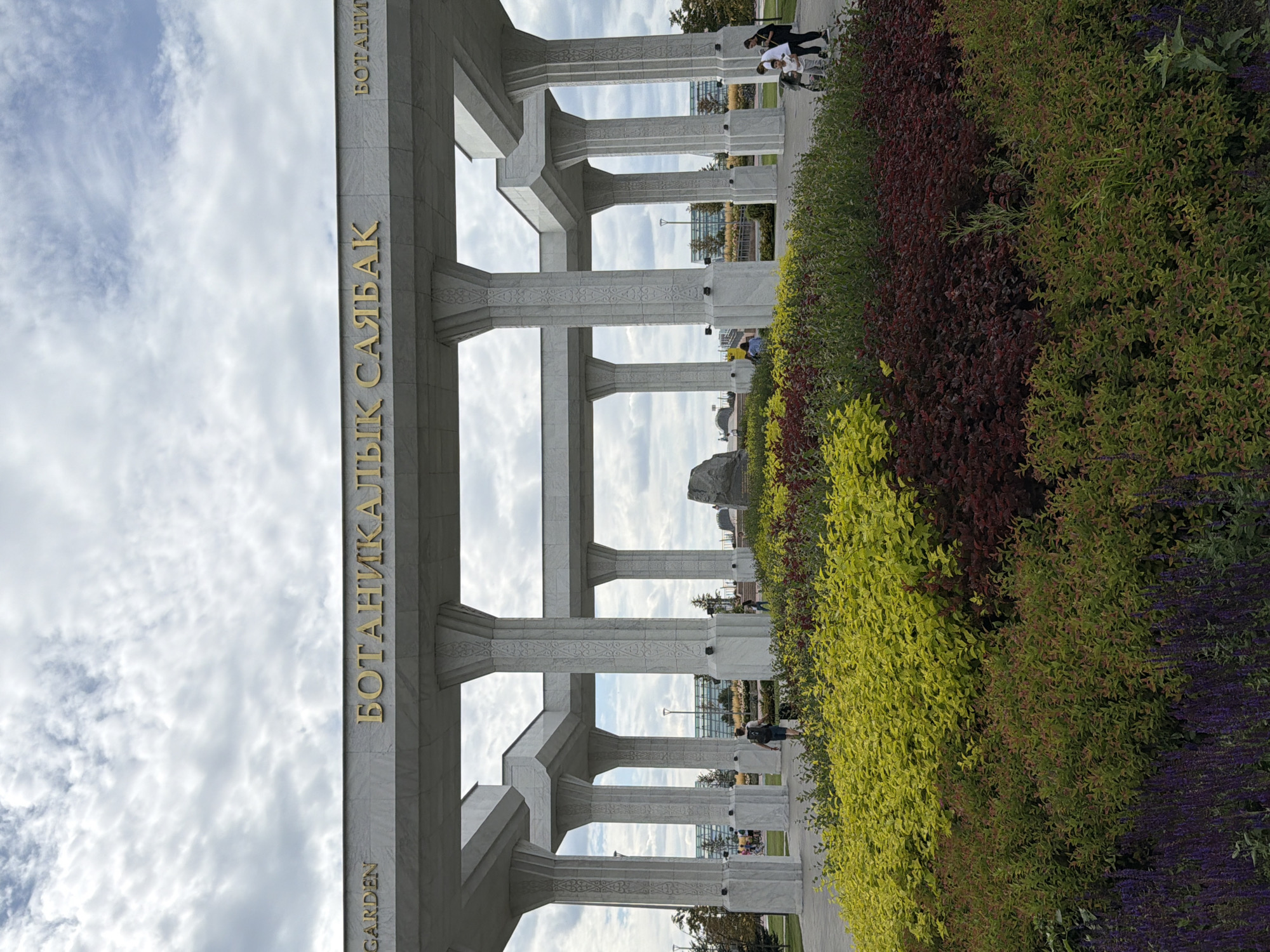 The image depicts the entrance to the Botanical Garden in Astana, Kazakhstan. A large, ornate marble structure with multiple pillars frames the view. Gold lettering on the structure reads "BOTANIKALYK SAYBAQ" in the center, with "GARDEN" on the left and "БОТАНИ" (likely the beginning of "БОТАНИЧЕСКИЙ") on the right. In the foreground, a colorful arrangement of plants features vibrant yellow bushes, deep red shrubs, and purple flowers. Several people are visible walking on a paved path behind the plants and under the archway. The sky is overcast with scattered clouds.