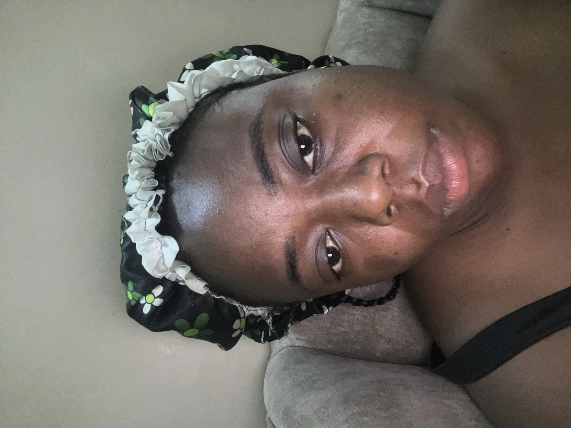 A close-up, horizontal shot captures a person of color, appearing to be a woman, lying down and looking directly at the camera. Her head is positioned towards the left of the frame, and her body extends to the right. She has dark skin with a visible sheen, particularly on her forehead and cheeks. Her eyes are open, and her lips are slightly parted in a neutral expression.
She is wearing a dark-colored sleep bonnet or satin cap adorned with a pattern of white and light green flowers, and a ruffled white elastic trim around the edge. A dark, possibly braided, strand of hair is visible near her right ear. Part of a dark strap, indicative of clothing like a bra or camisole, is visible near her shoulder.
The background consists of a plain, light-colored surface on the left, possibly a wall, and a textured grey upholstered surface on the right, which appears to be a couch or bed. The overall setting is indoors. There are no visible textual elements in the image. The general lighting suggests it could be daytime or well-lit indoors. The provided context for the image is Abuja, Nigeria, although there are no explicit visual cues within the image itself to confirm this location.