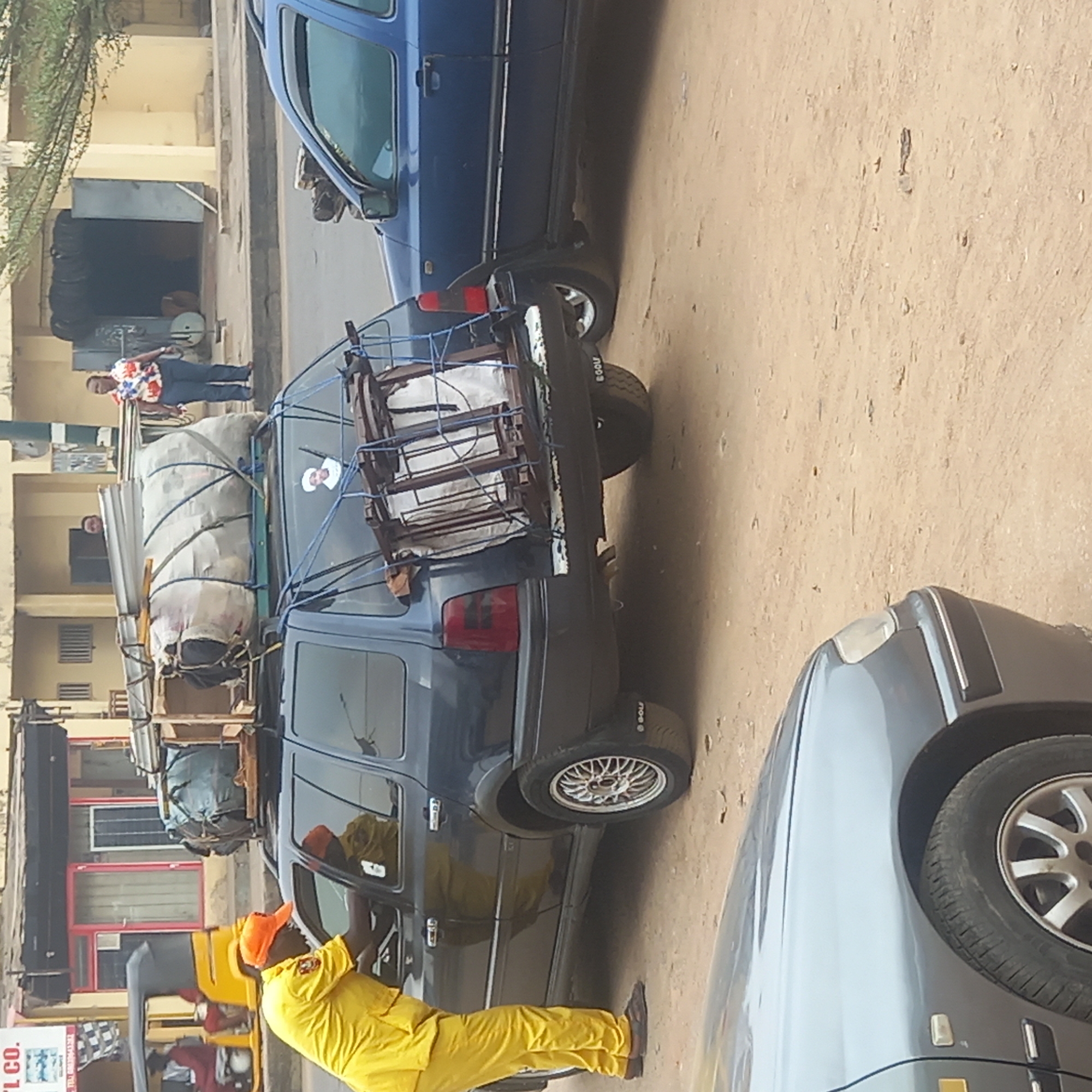 The image shows a street scene in Dadi Riba, Nigeria. A dark blue station wagon is heavily loaded with items strapped to its roof, including bags and a metal frame. A man in a bright yellow uniform and orange cap stands next to the car, appearing to interact with it. Two other vehicles, one blue and one gray, are partially visible in the frame. In the background, a man in a colorful shirt stands on the sidewalk next to some buildings. The street surface is dusty.