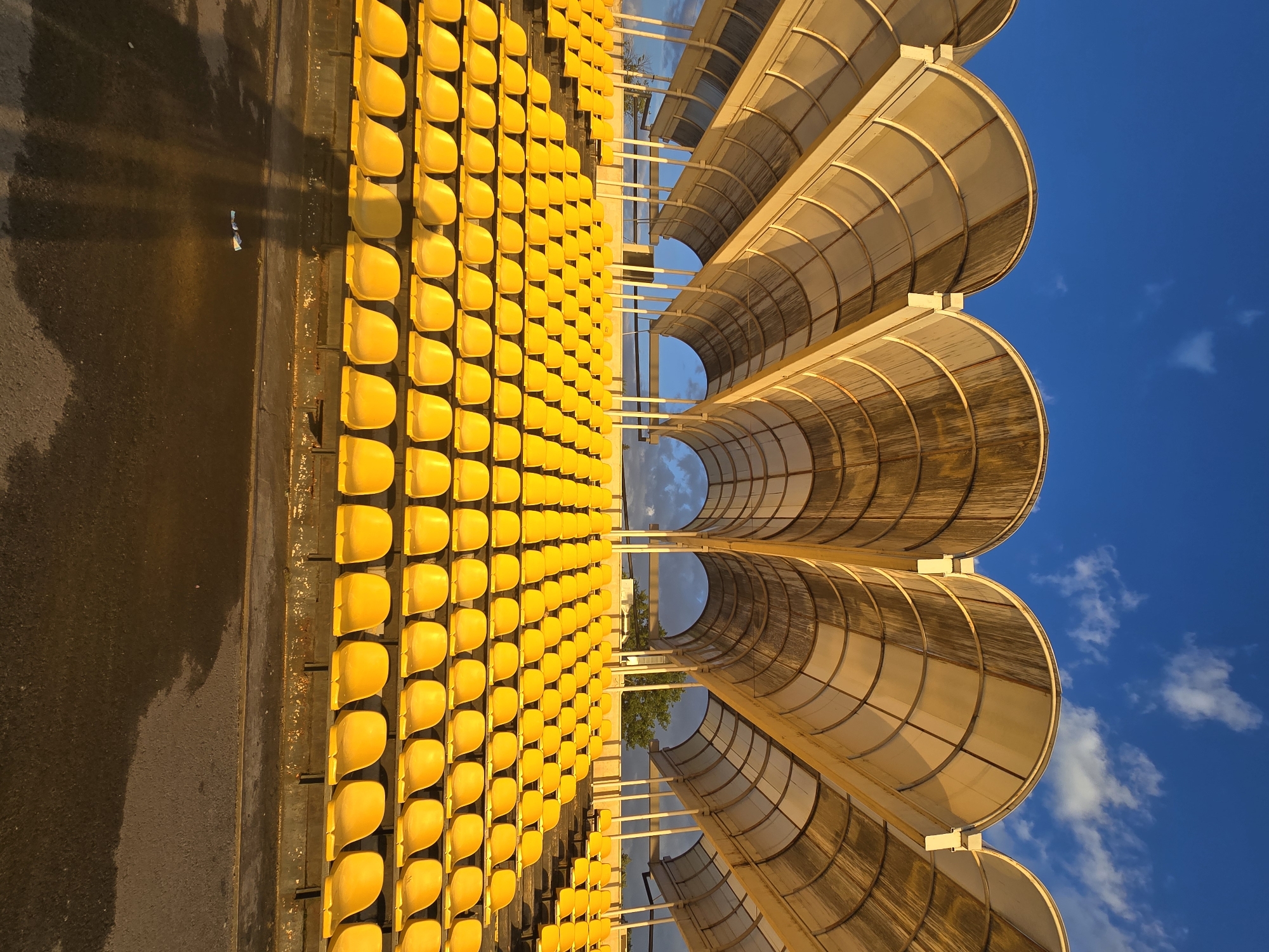 The image shows a partial view of a stadium with rows of yellow seats on the left side and a curved, covered structure on the right, against a blue sky with some clouds. The seats are arranged in an orderly manner, and the covered structure appears to provide shade or protection. The foreground includes a paved area, possibly a walkway or access point to the seating area. The photograph is taken from an elevated perspective, offering a clear view of the stadium's architecture and seating arrangement. The specific location within the city of Unknown, Unknown, cannot be determined from the given image.