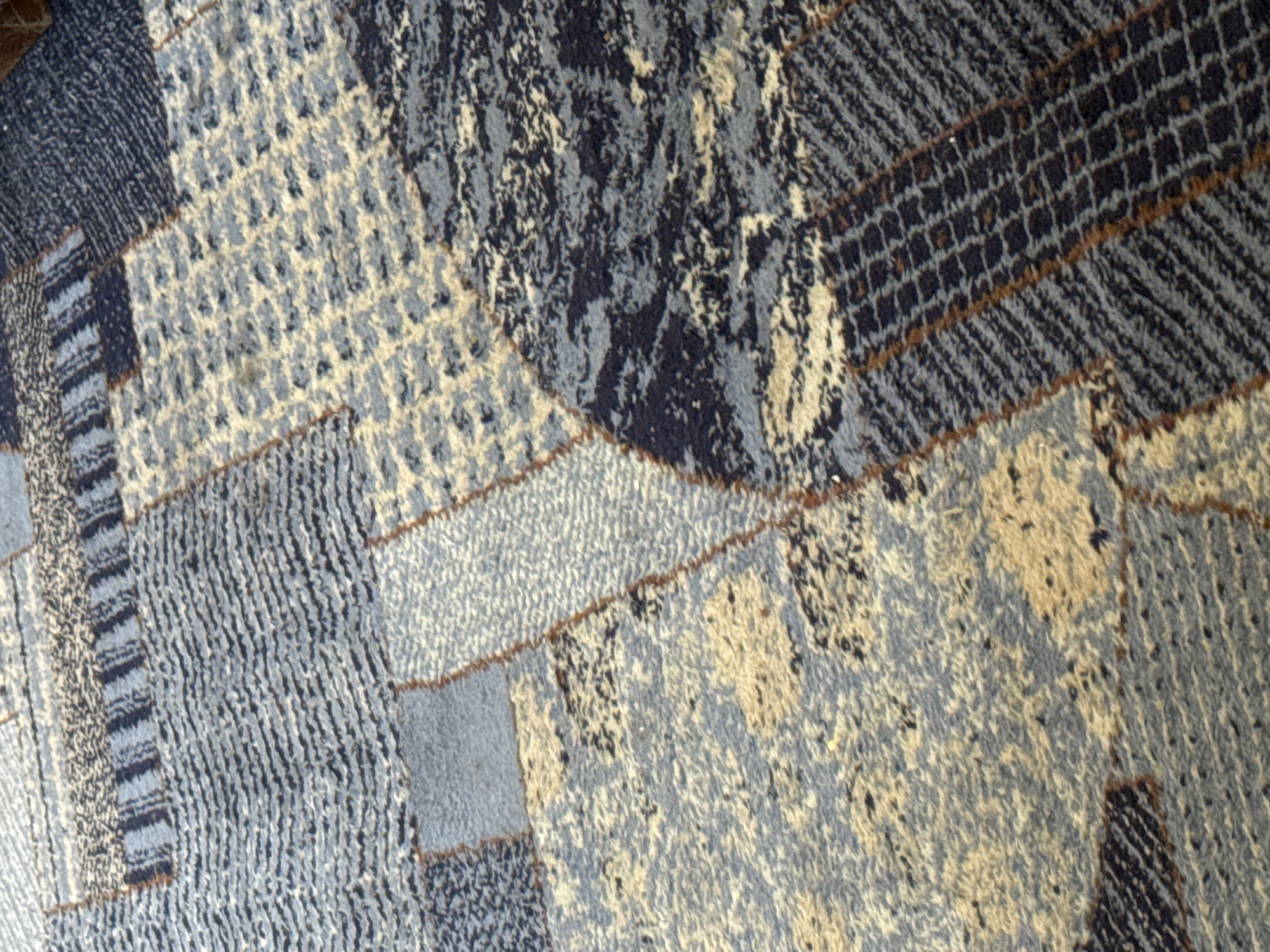 The image displays a close-up, overhead view of a carpet with a geometric, abstract pattern. The carpet features a patchwork of rectangular and irregularly shaped sections in various shades of blue, beige, and dark gray. Some sections have a ribbed texture, while others have a more granular or speckled appearance. Thin brown lines or threads are visible, outlining some of the shapes and adding to the intricate design. The lighting suggests an indoor environment, likely a room with carpeted flooring, though no specific location within Bishkek City, Kyrgyzstan is identifiable from the visual data provided. There are no people or discernible objects other than the carpet itself.